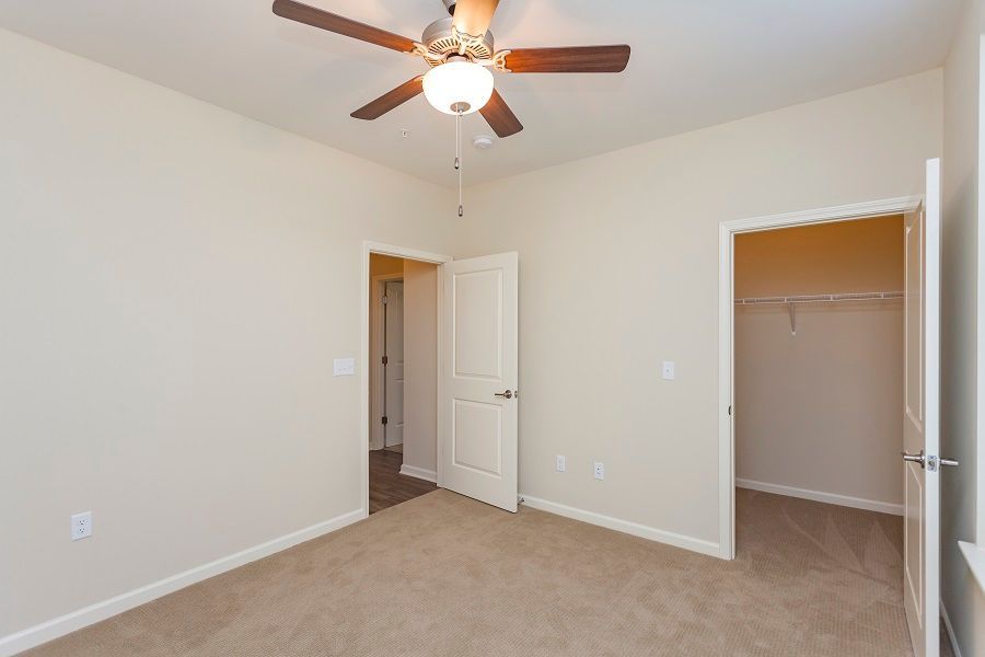 An empty room with beige carpet and walls, a ceiling fan, a partially open door, and a walk-in closet.