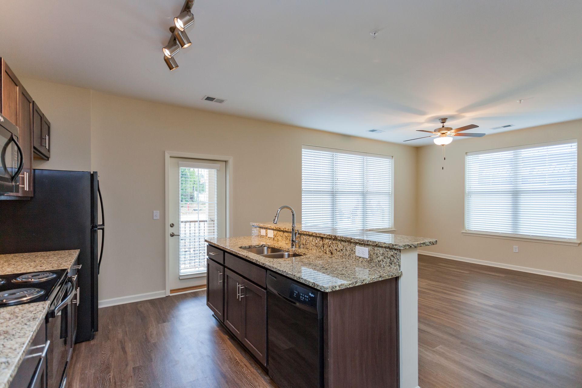 A kitchen with brown cabinets, granite countertops, and black appliances opens into a spacious, sunlit living area.