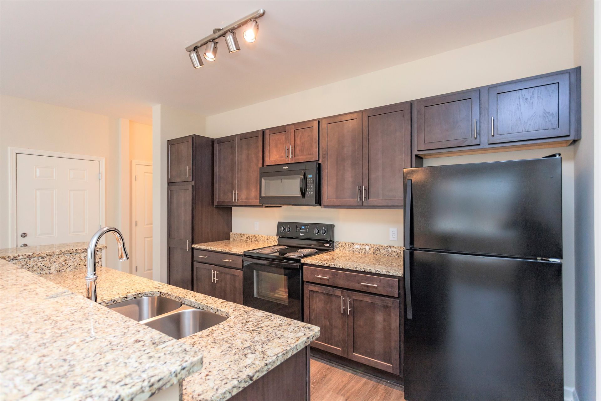 A modern kitchen featuring dark wood cabinets, speckled granite countertops, stainless steel sink, and black appliances.