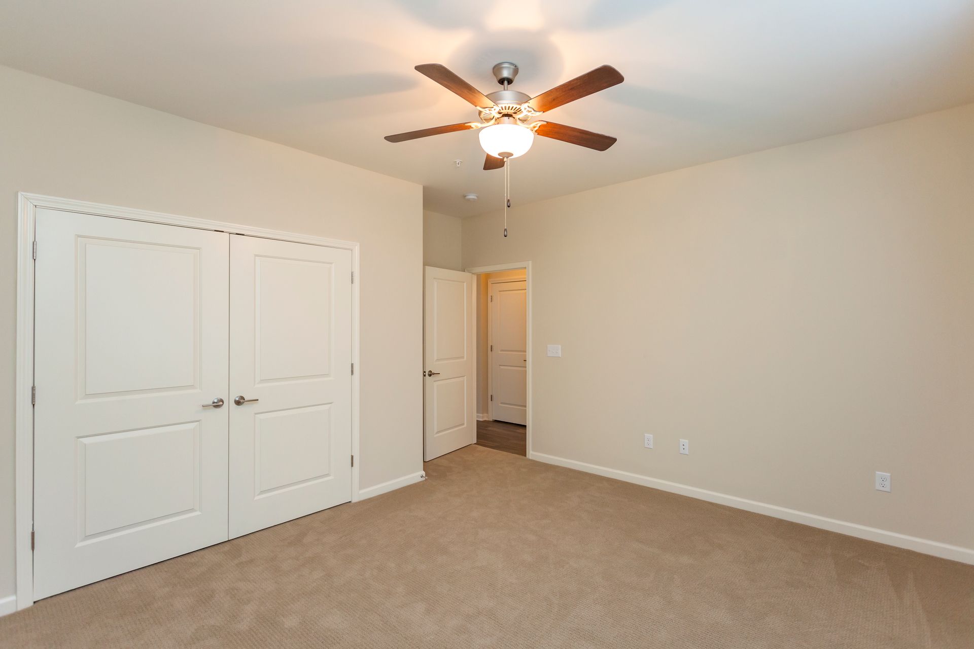 An empty bedroom with tan carpet, beige walls, a ceiling fan, and white doors leading to a closet and hallway.