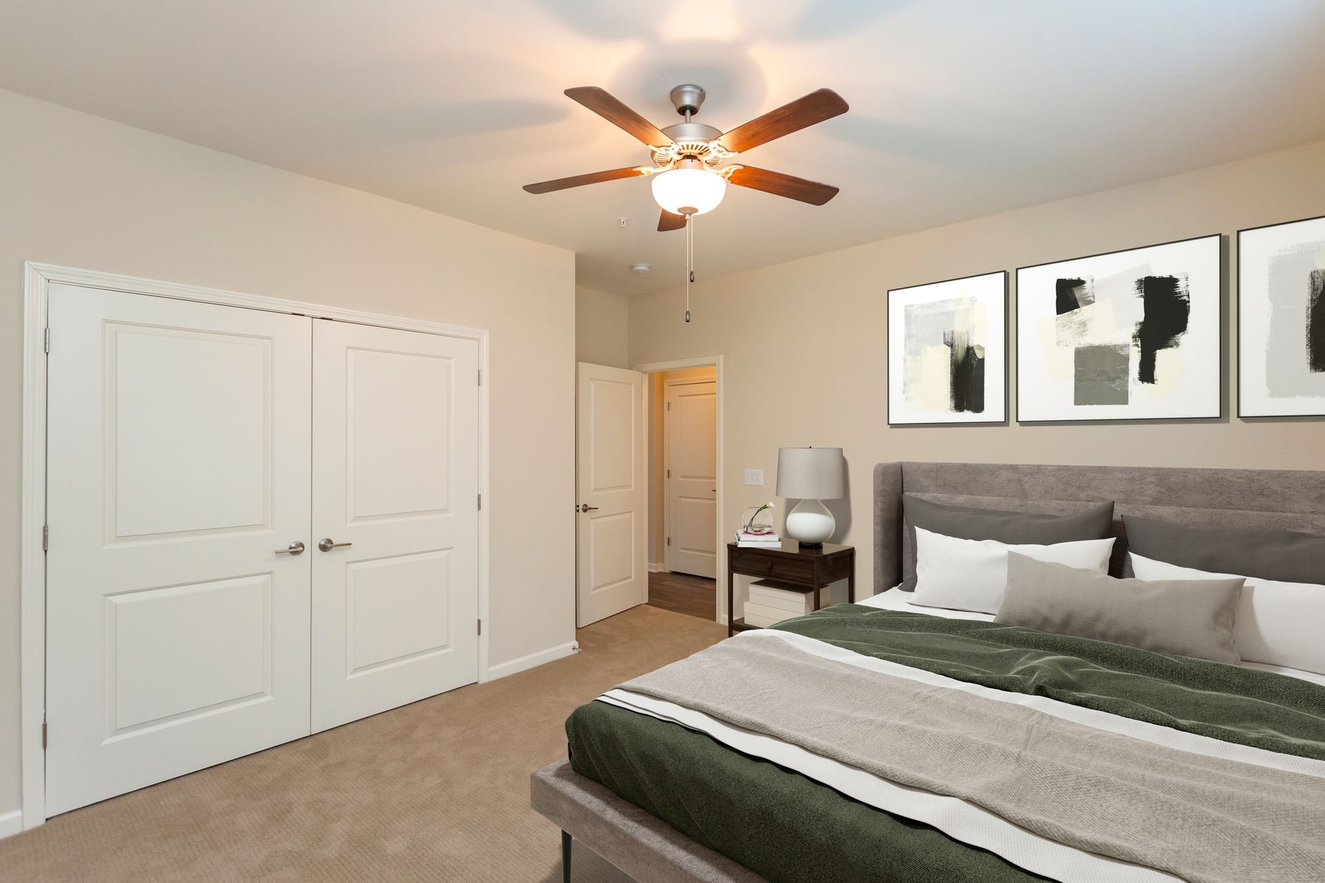 A modern bedroom with a large bed, double doors, a ceiling fan, and framed abstract art on a beige wall.