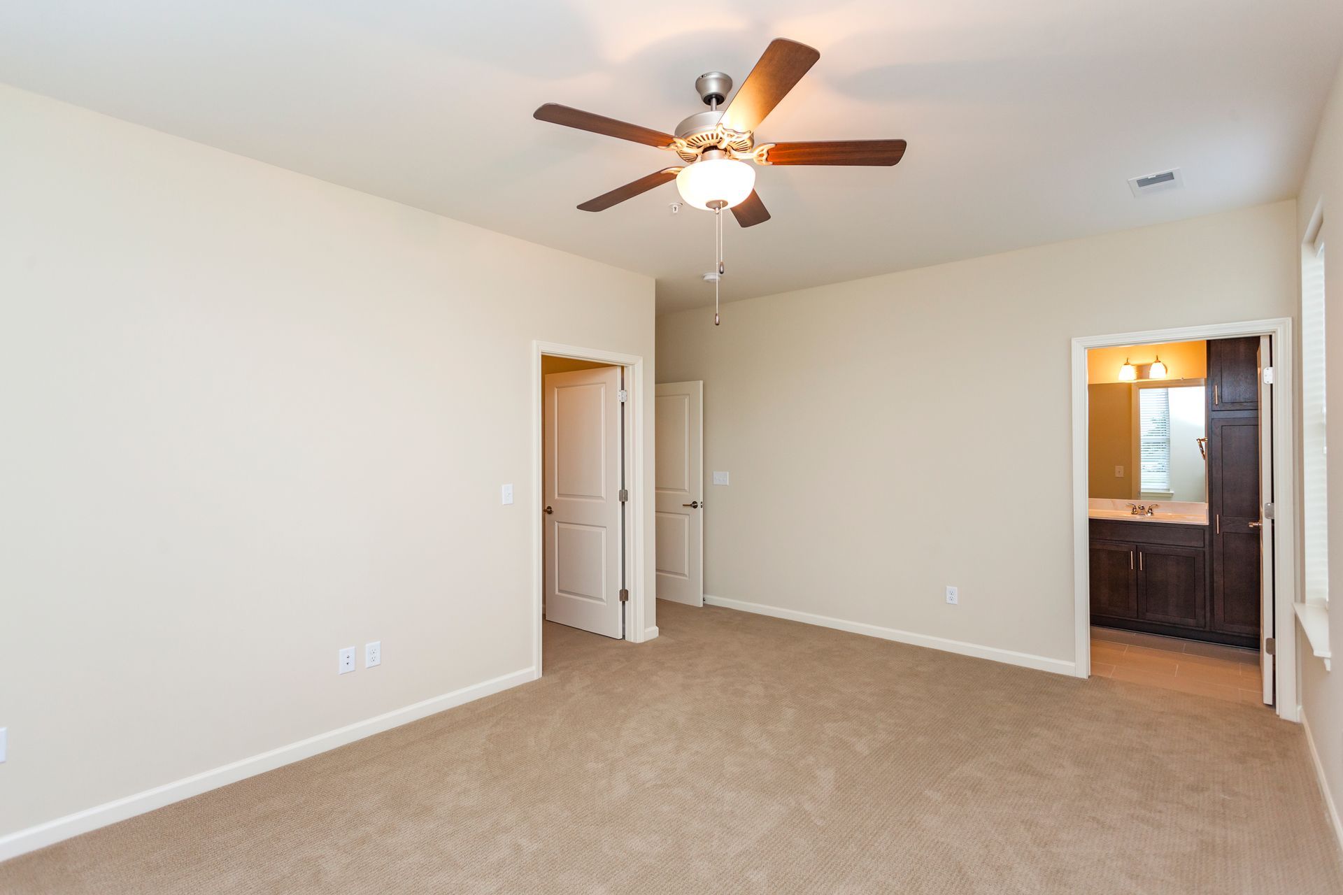 A spacious, empty bedroom with beige walls, tan carpeting, a ceiling fan, and doors leading to a closet and a bathroom.