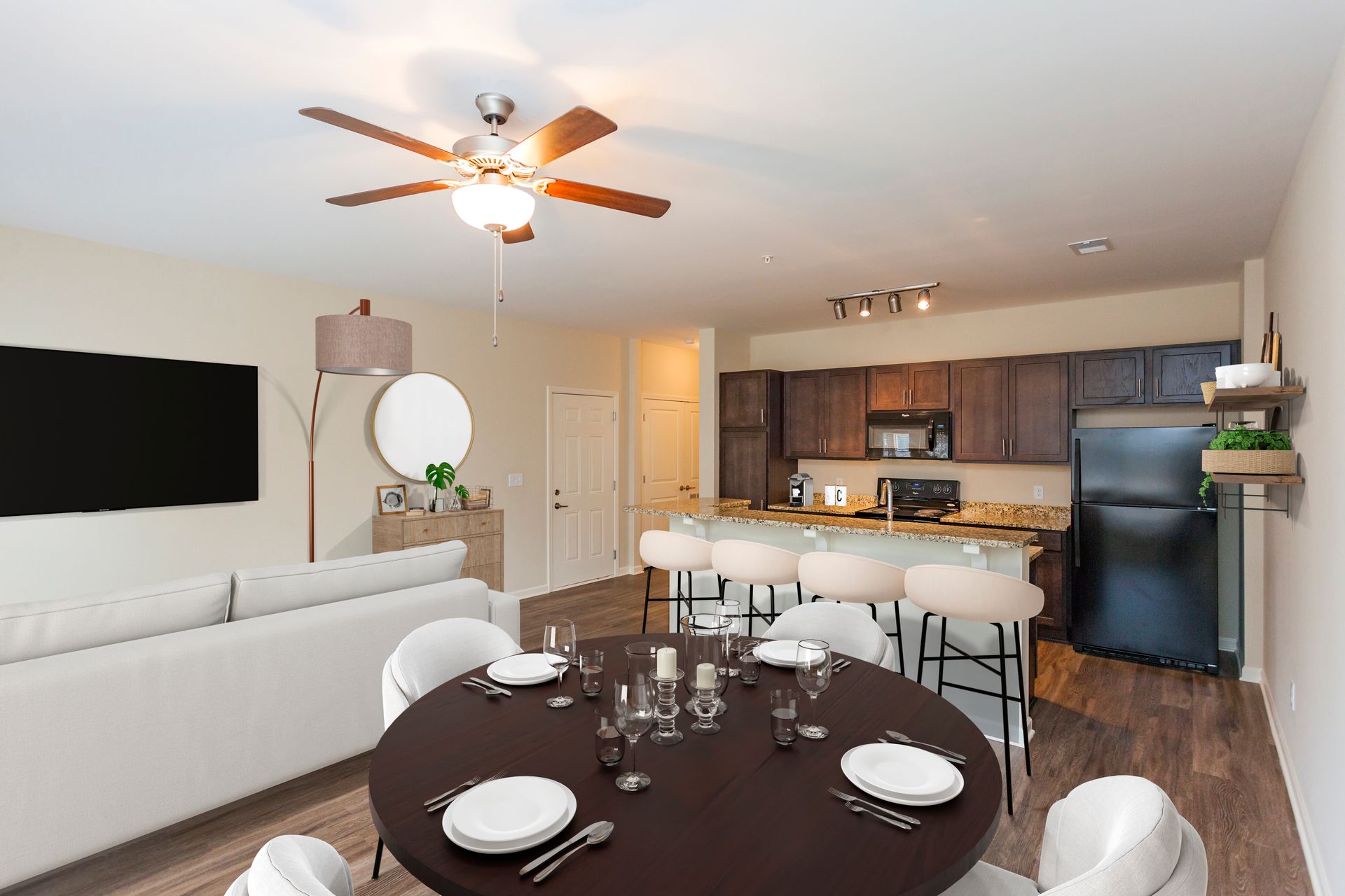 An open-concept living area with a round dining table, white sofa, and kitchen with dark wood cabinets and a black fridge.