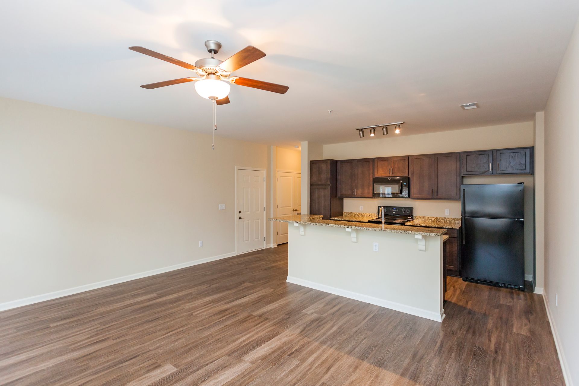 An open-concept room with wood-look flooring, a ceiling fan, and a kitchen featuring dark cabinets and a black fridge.