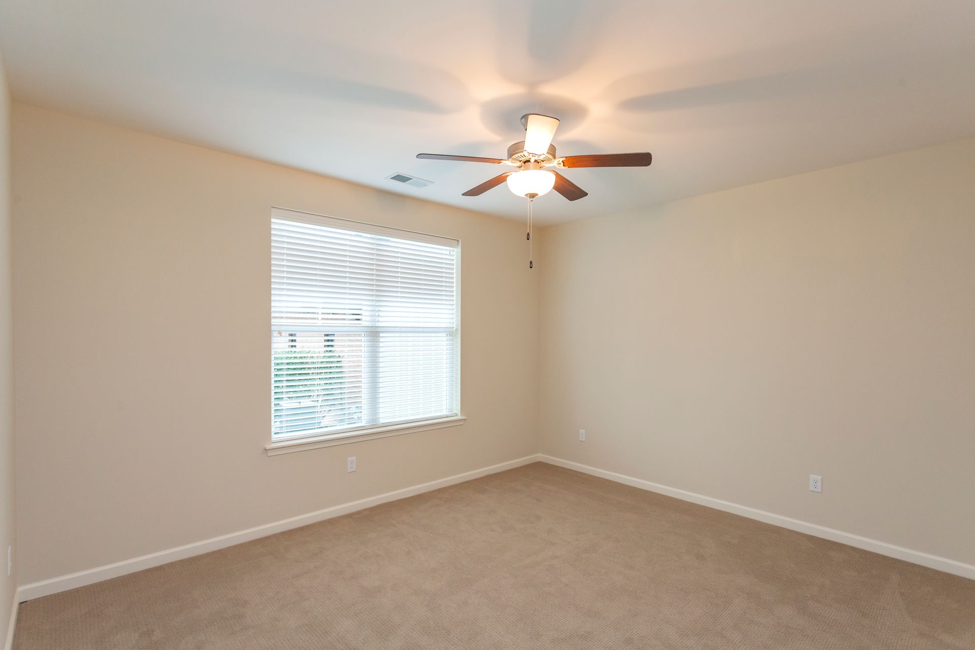 An empty, neutrally painted bedroom with beige carpet, a single window with blinds, and a ceiling fan with light.