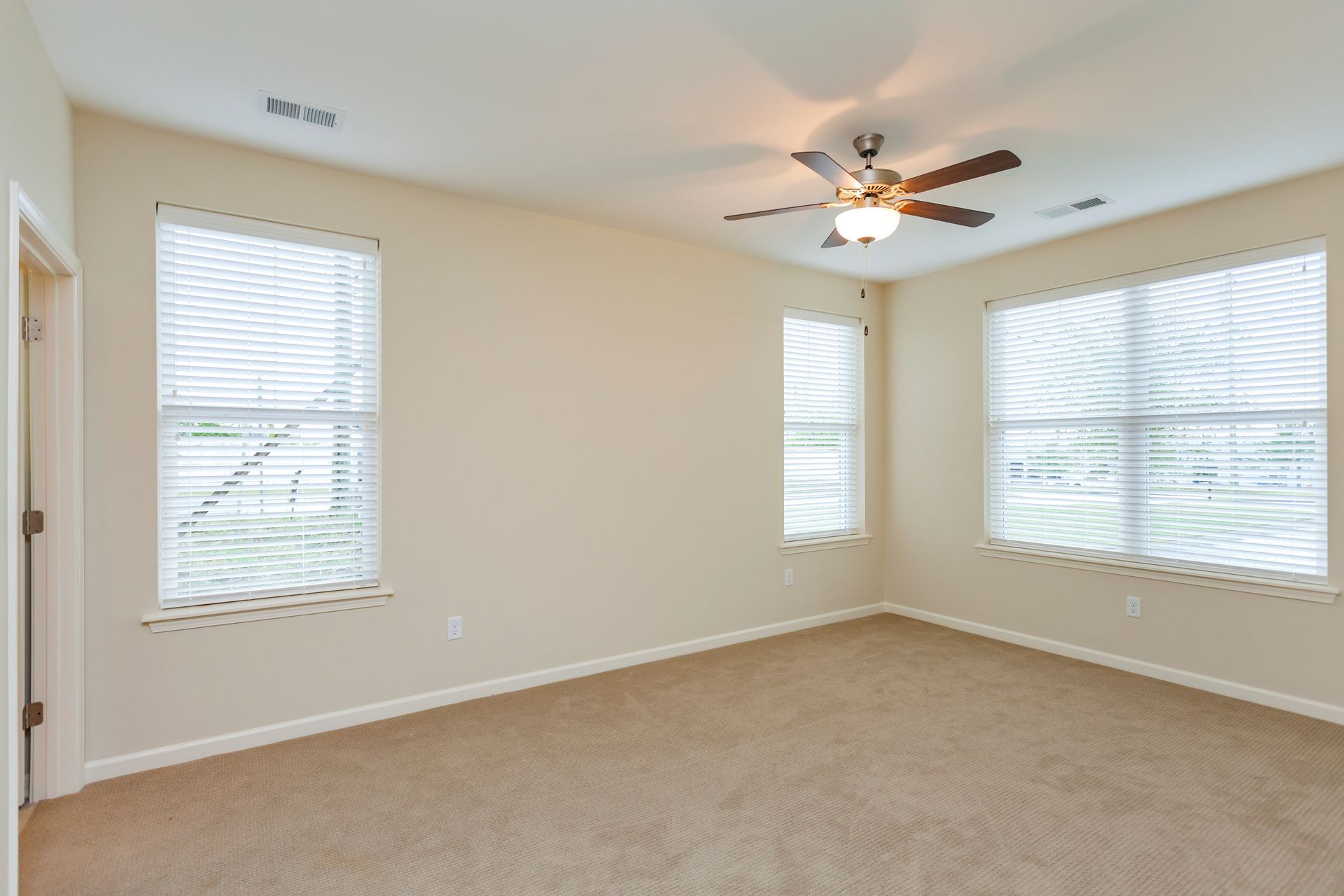 Empty room with tan carpet and walls, white trim, a ceiling fan, and three windows covered with white horizontal blinds.