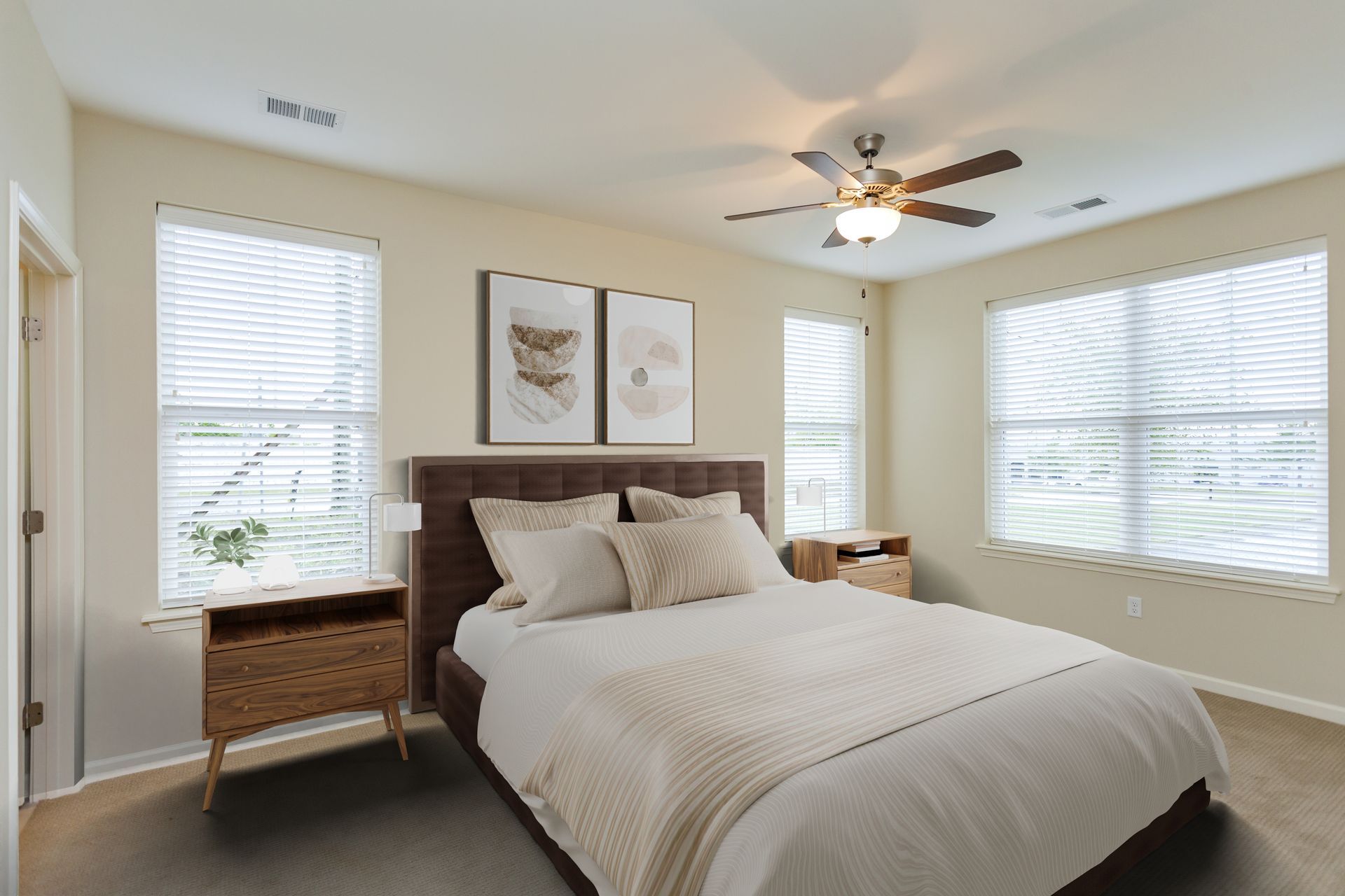 A bedroom with a brown upholstered headboard, a white bed, a wooden nightstand, and two framed prints on a cream wall.