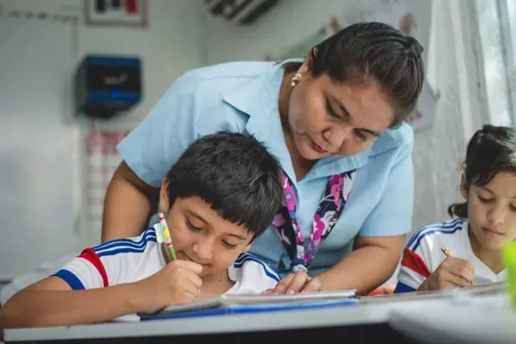 Profesora ayudando a un estudiante a escribir en un escritorio; otro estudiante cerca.
