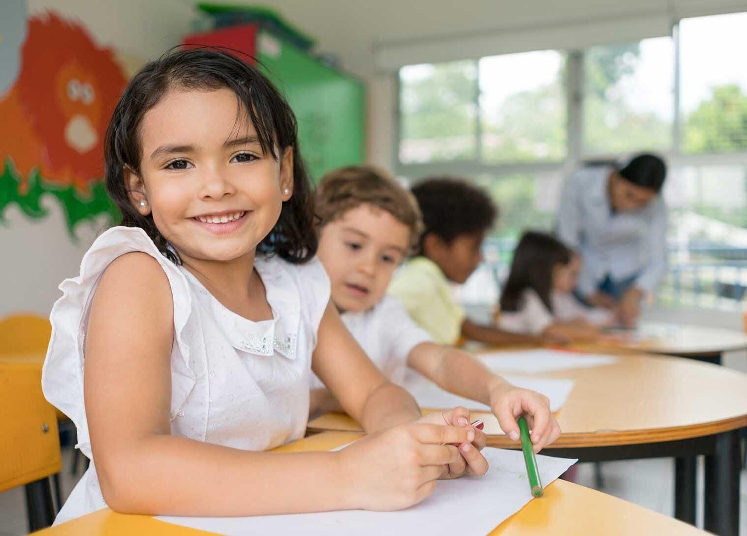 Niño sonriente en un escritorio en un aula, otros niños y un maestro en el fondo.