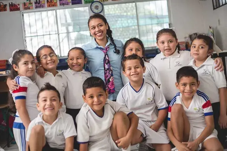 La maestra posa con un grupo sonriente de estudiantes en un aula bien iluminada.