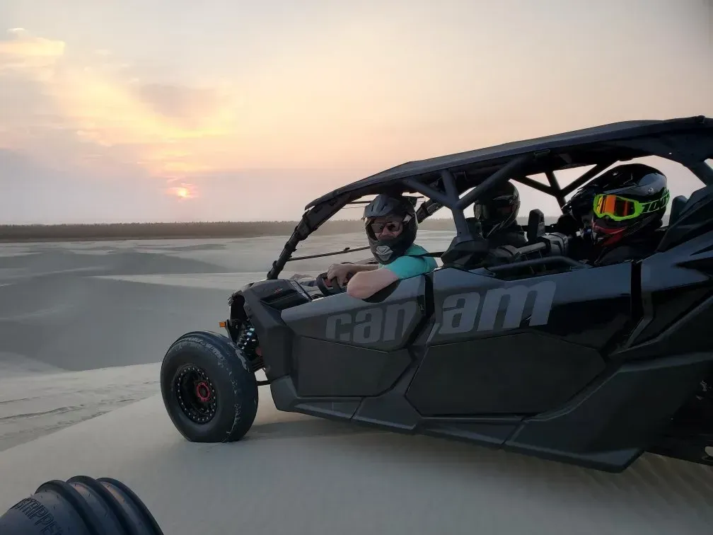 Two people wearing helmets ride a dune buggy across sand dunes at sunset. The open vehicle is labeled 