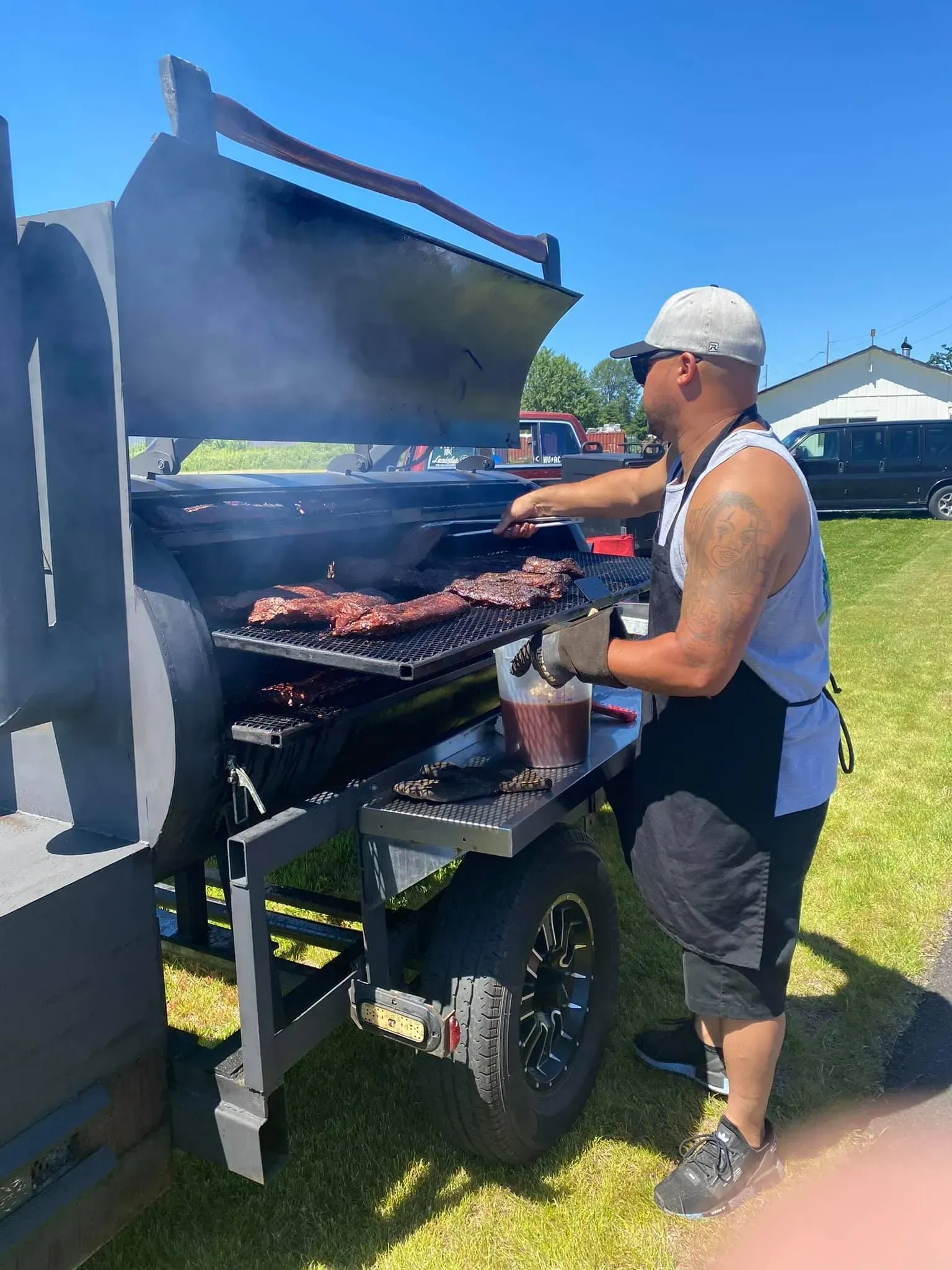 A man wearing a white cap and apron is grilling ribs on a large outdoor barbecue smoker.