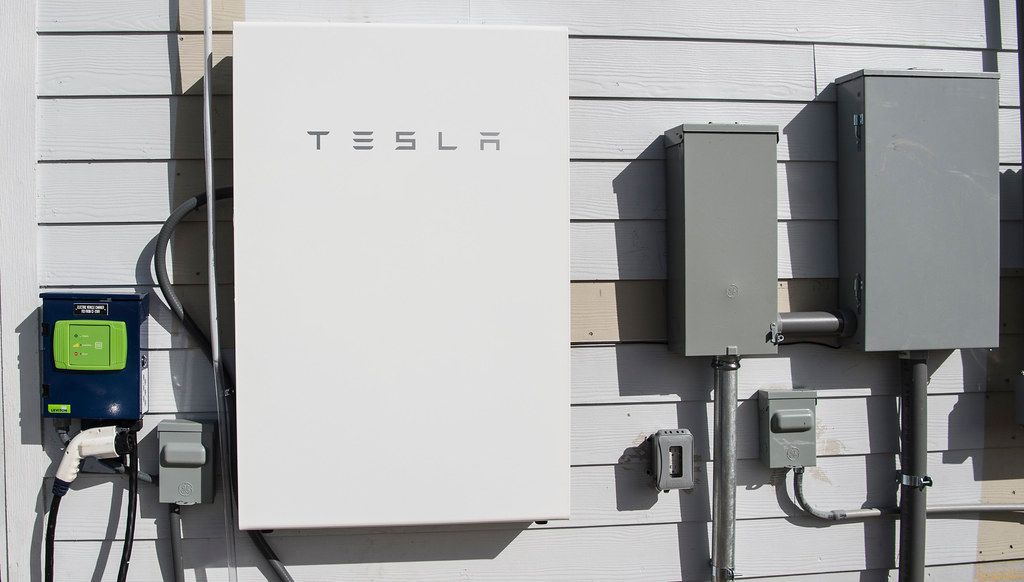 A Tesla Powerwall is mounted on a wall next to other electrical panels and devices. The setup is part of a home energy system