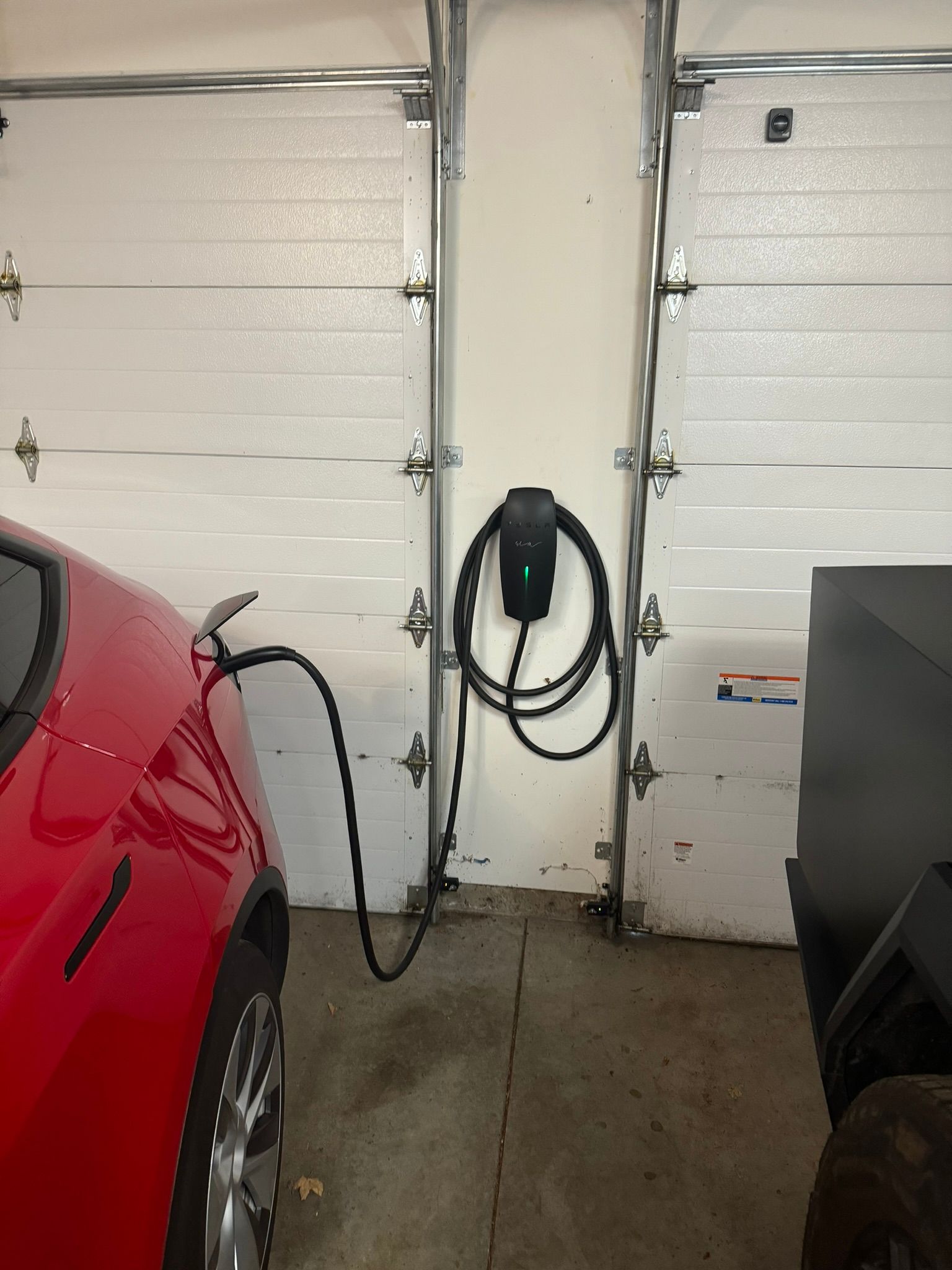 Red car parked in garage connected to wall-mounted charging station with black cable.