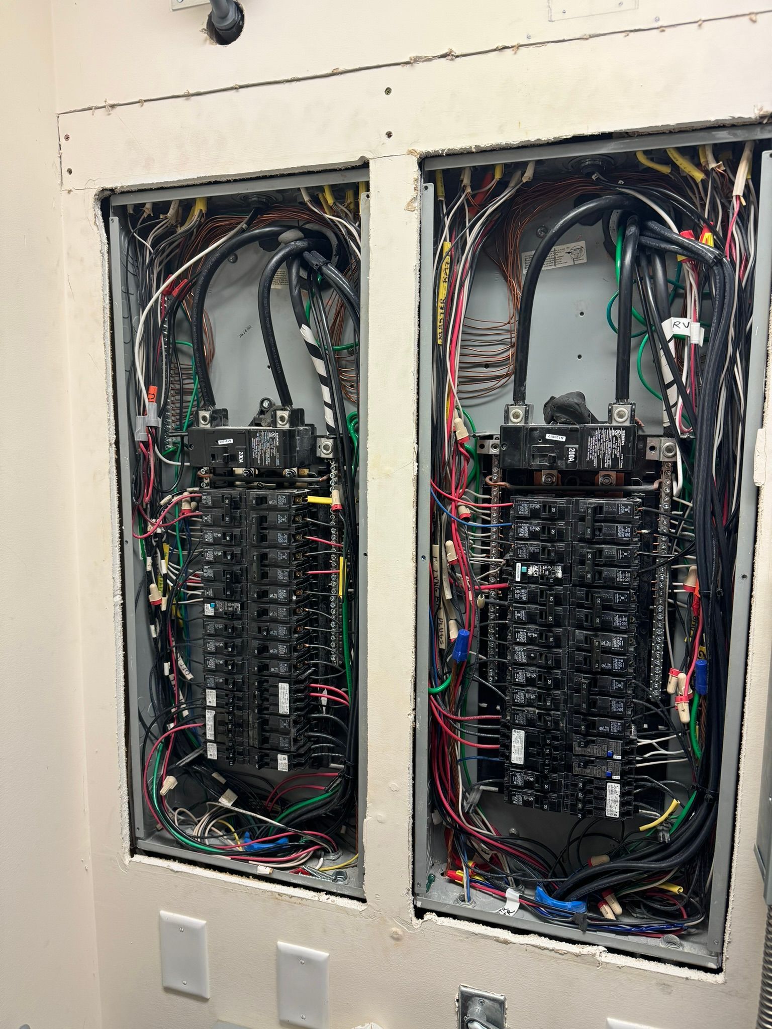 Two open electrical panels with numerous wires of different colors connected to circuit breakers inside a wall.
