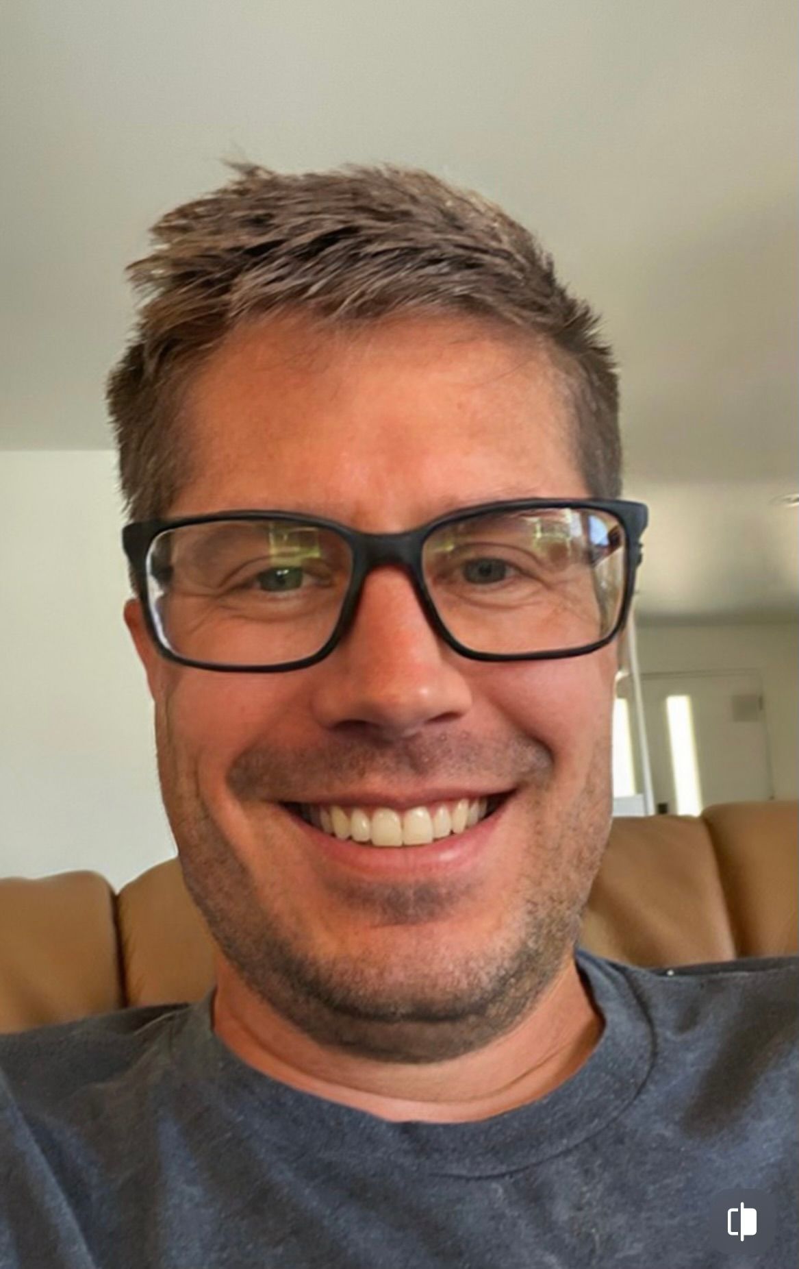 A man with short brown hair and glasses is smiling while sitting indoors.