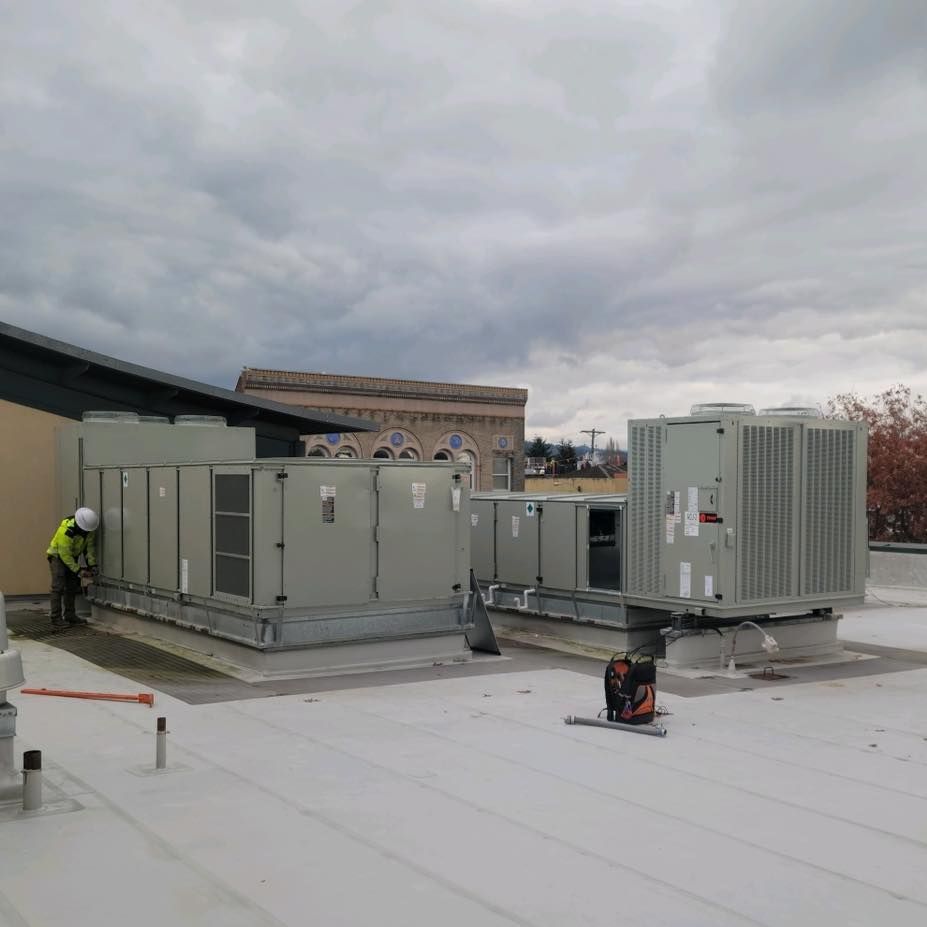 A worker in safety gear inspects large HVAC units on a flat rooftop under a cloudy sky.