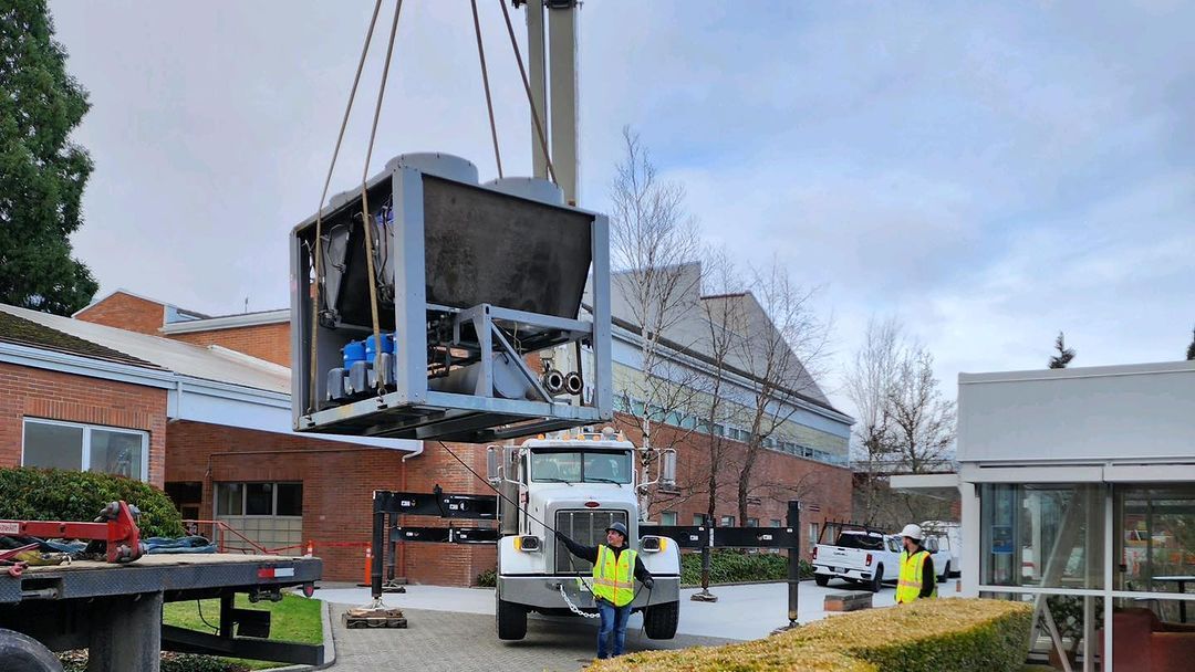 Crane lifting large industrial unit outdoors, two workers in yellow safety vests.