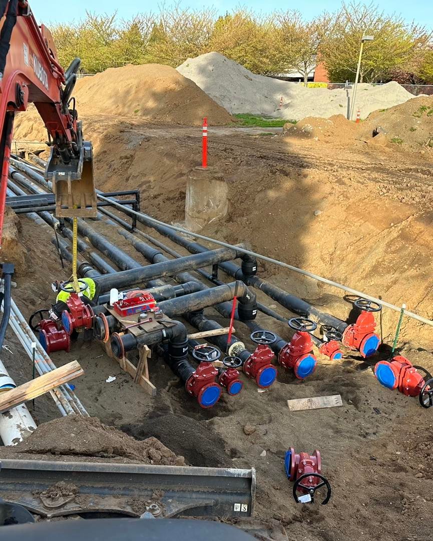 Construction site with large pipes and red valves in the trench, a workers in safety gear nearby.