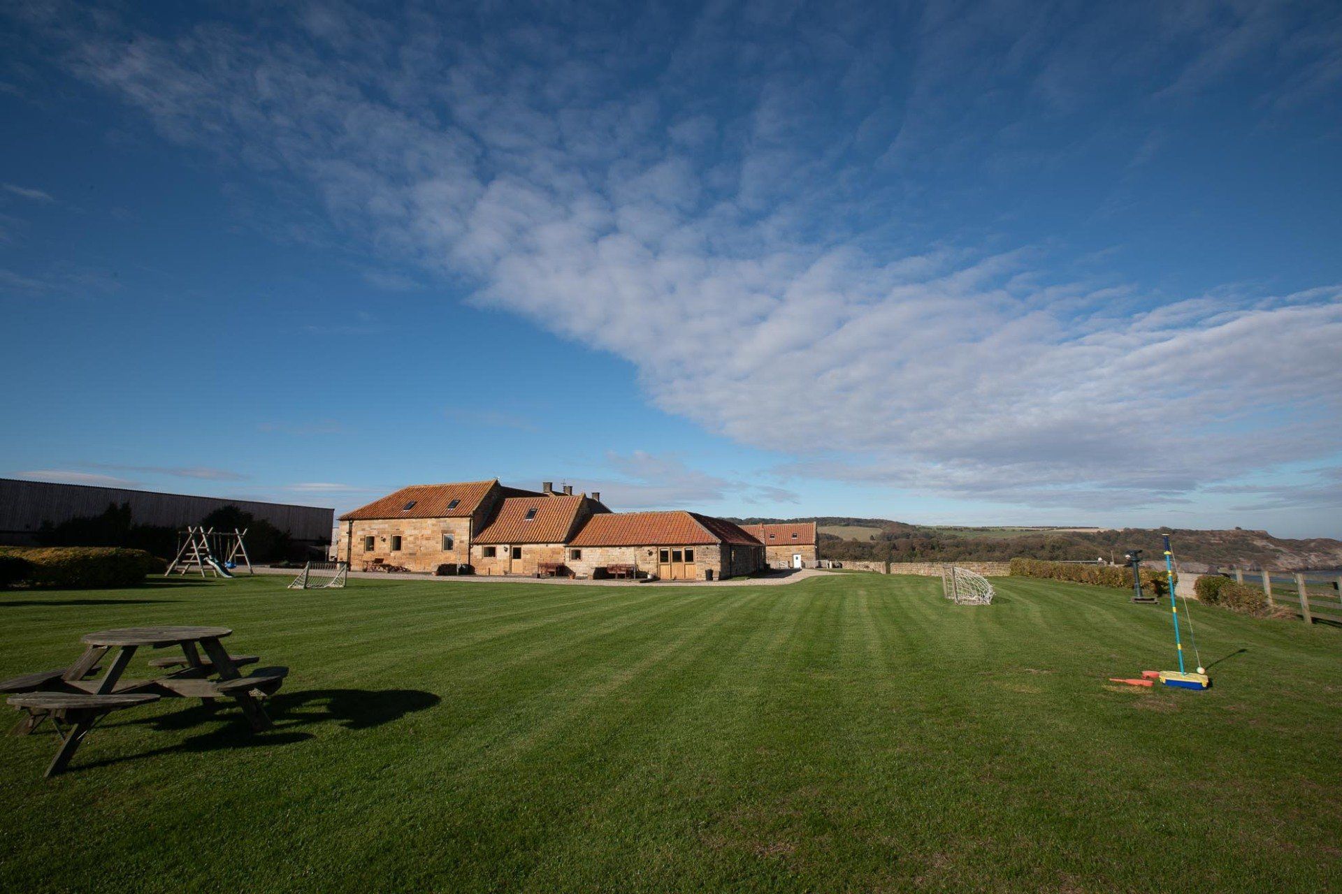 Lush green lawn with a large brick building under a blue sky with wispy clouds. Picnic table in the foreground.