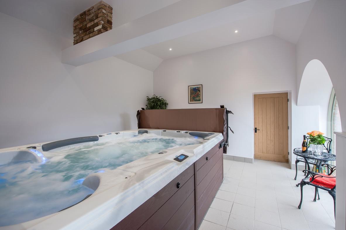 Indoor spa room with a long rectangular hot tub. White walls, wood door, and a small table.