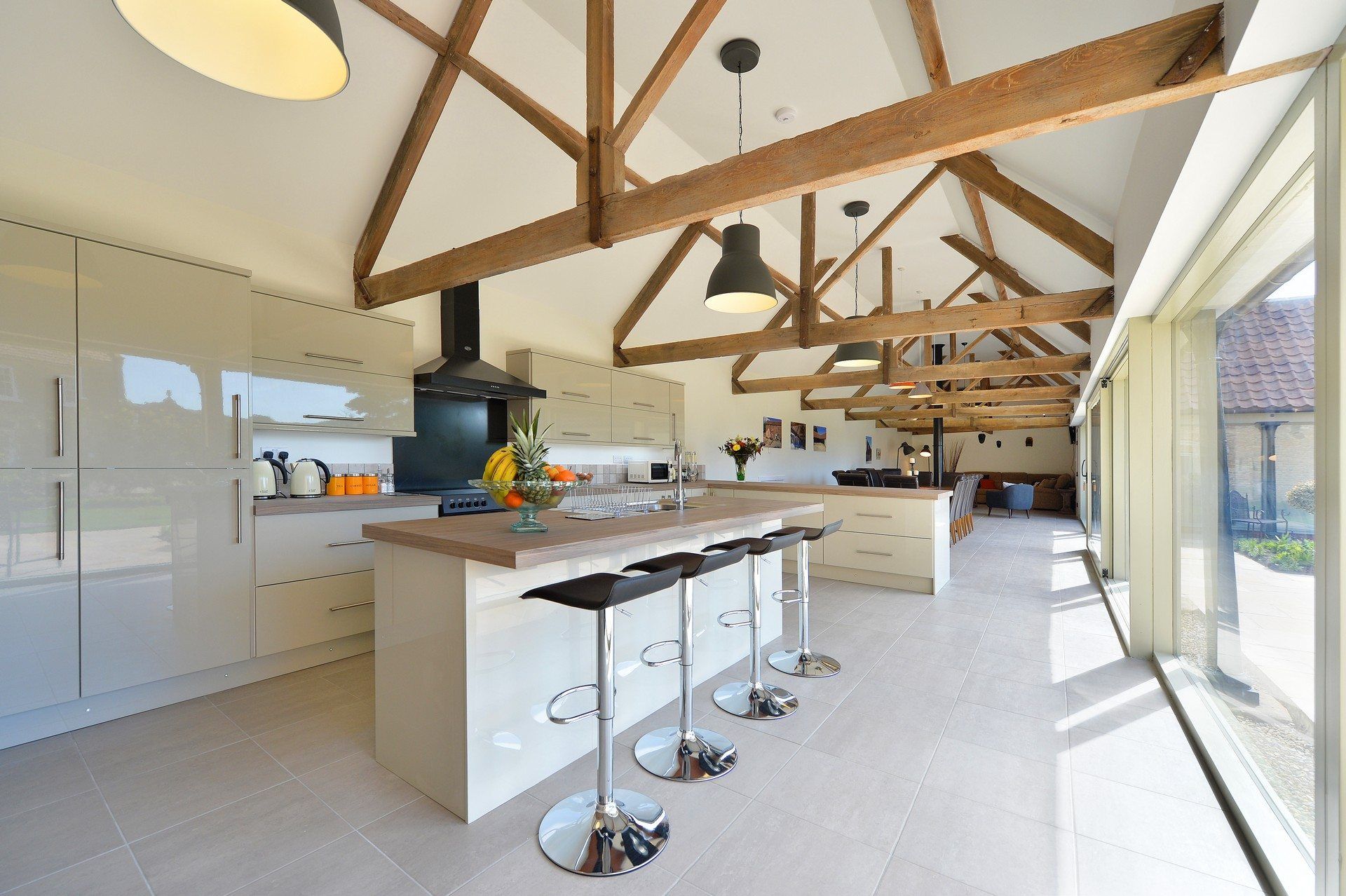 Modern kitchen with vaulted wooden beams, white cabinets, and island with bar stools. Large windows offer outside view.