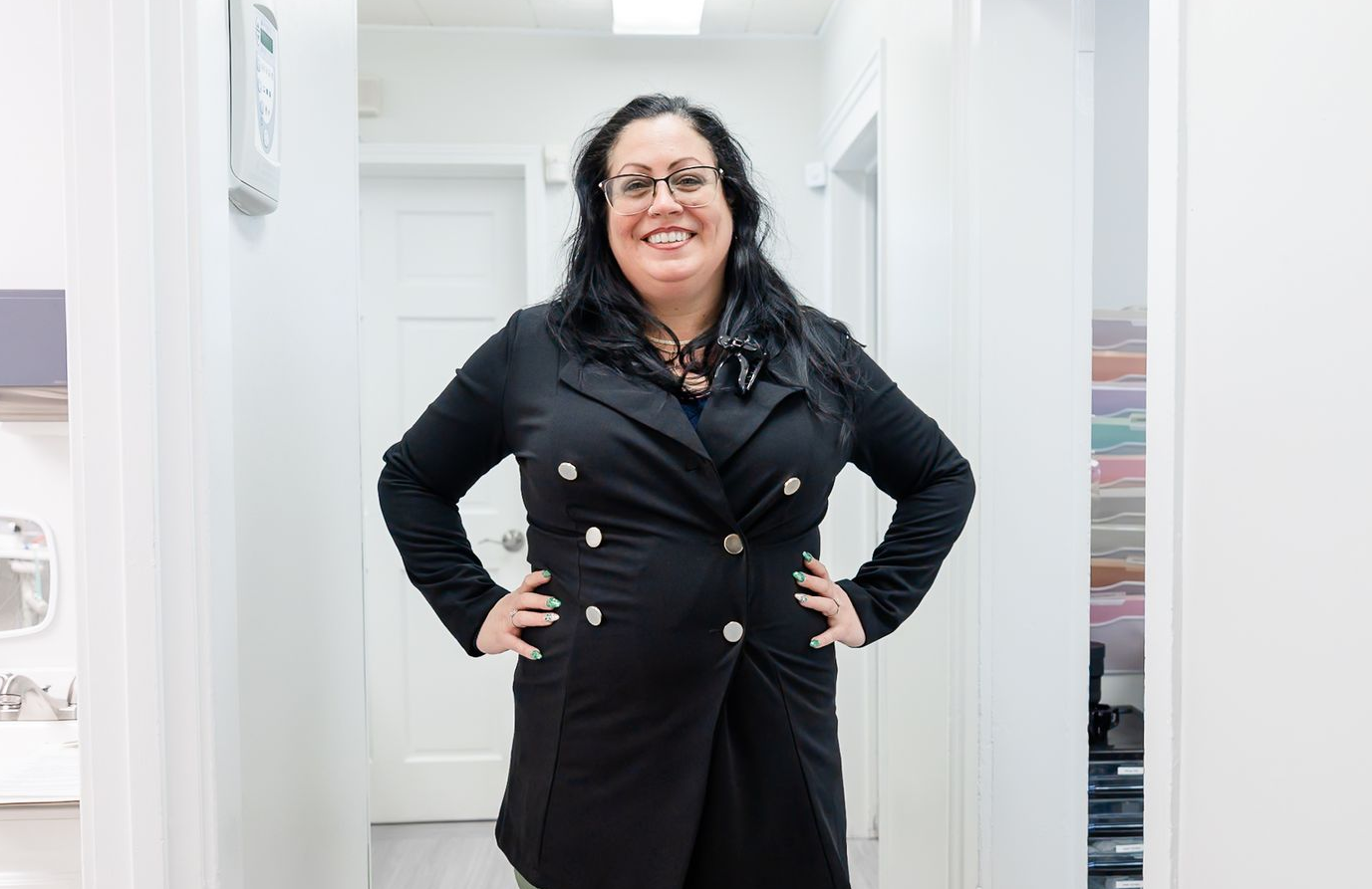Woman in black scrubs, hands on hips, smiling in a white hallway with a bright overhead light.