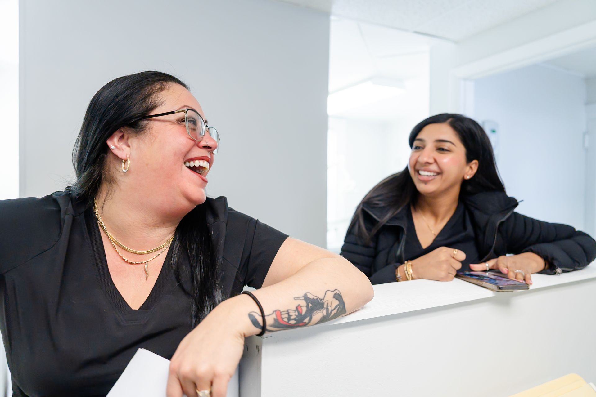 Two women laugh while standing behind a white reception desk in a brightly lit office.