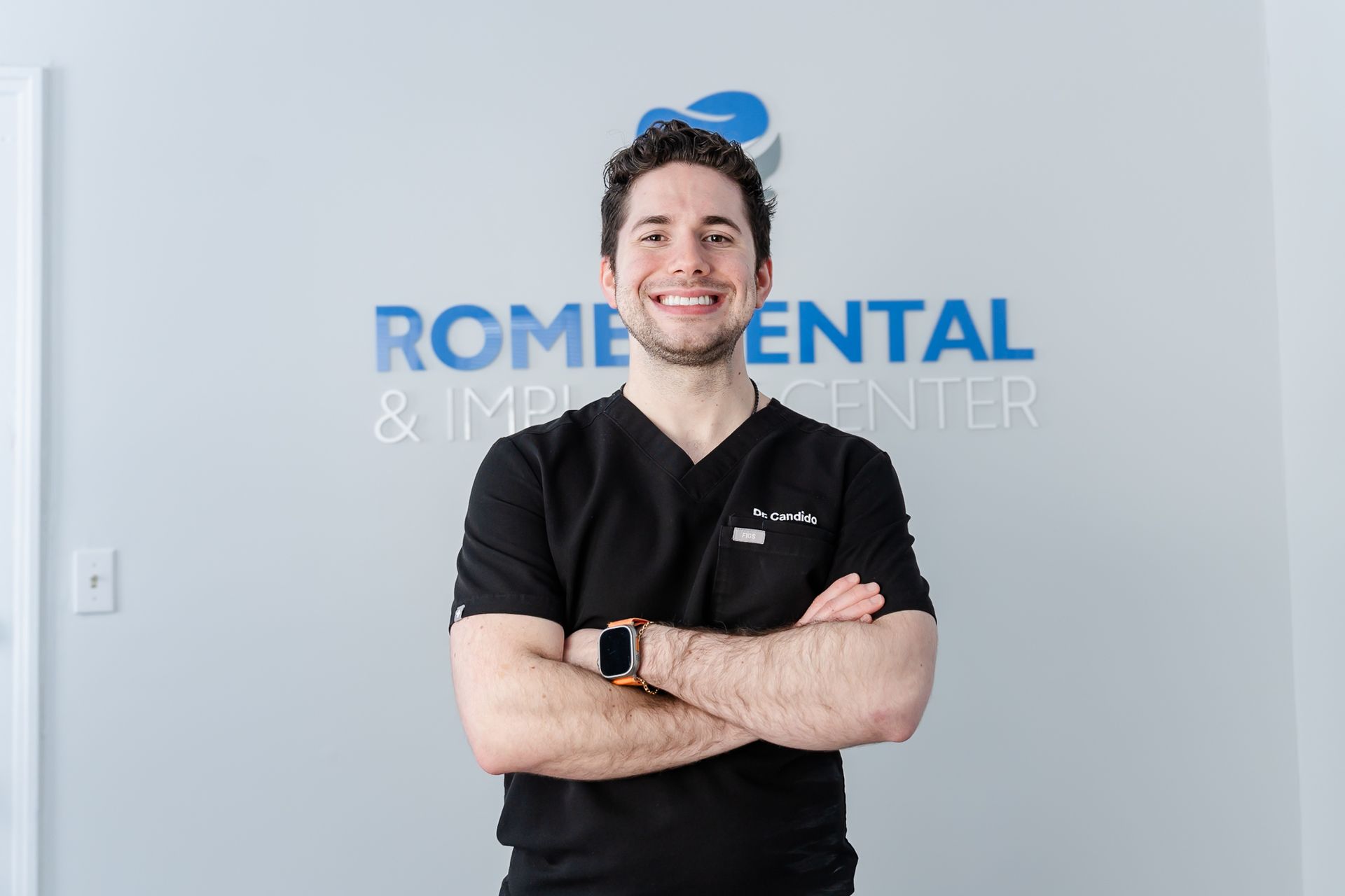 Man in black scrubs smiles with arms crossed in front of 