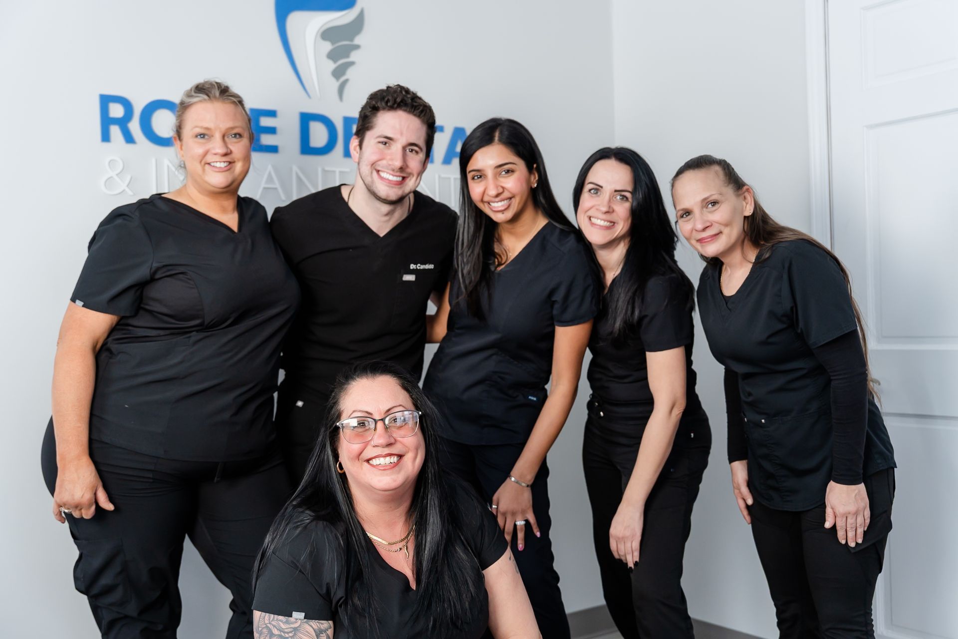 Group of six dental professionals smiling in front of a clinic logo. All are wearing black scrubs.