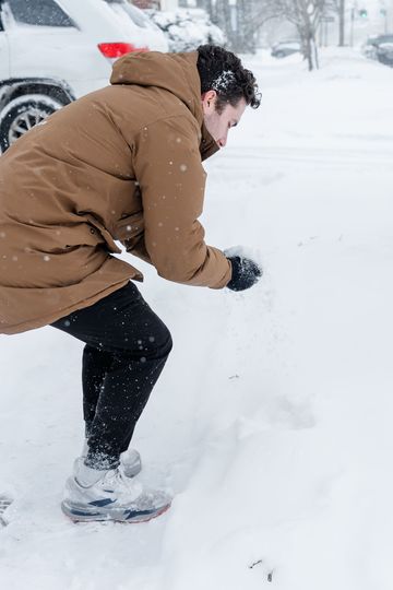 Man in brown jacket kneels in snow, forming snowball. Black pants, gloves, and white sneakers. Snowing.