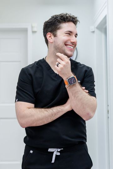 Man in black scrubs smiles, looking away; hands near face, watch visible. White-walled interior.