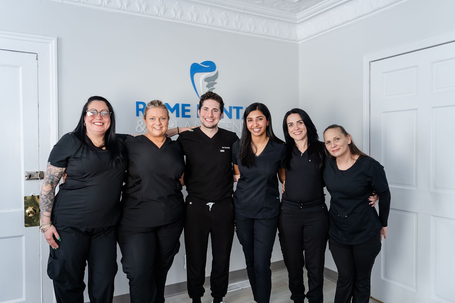 Dental office staff, six people, smiling, posing in front of logo. Everyone is wearing black scrubs.