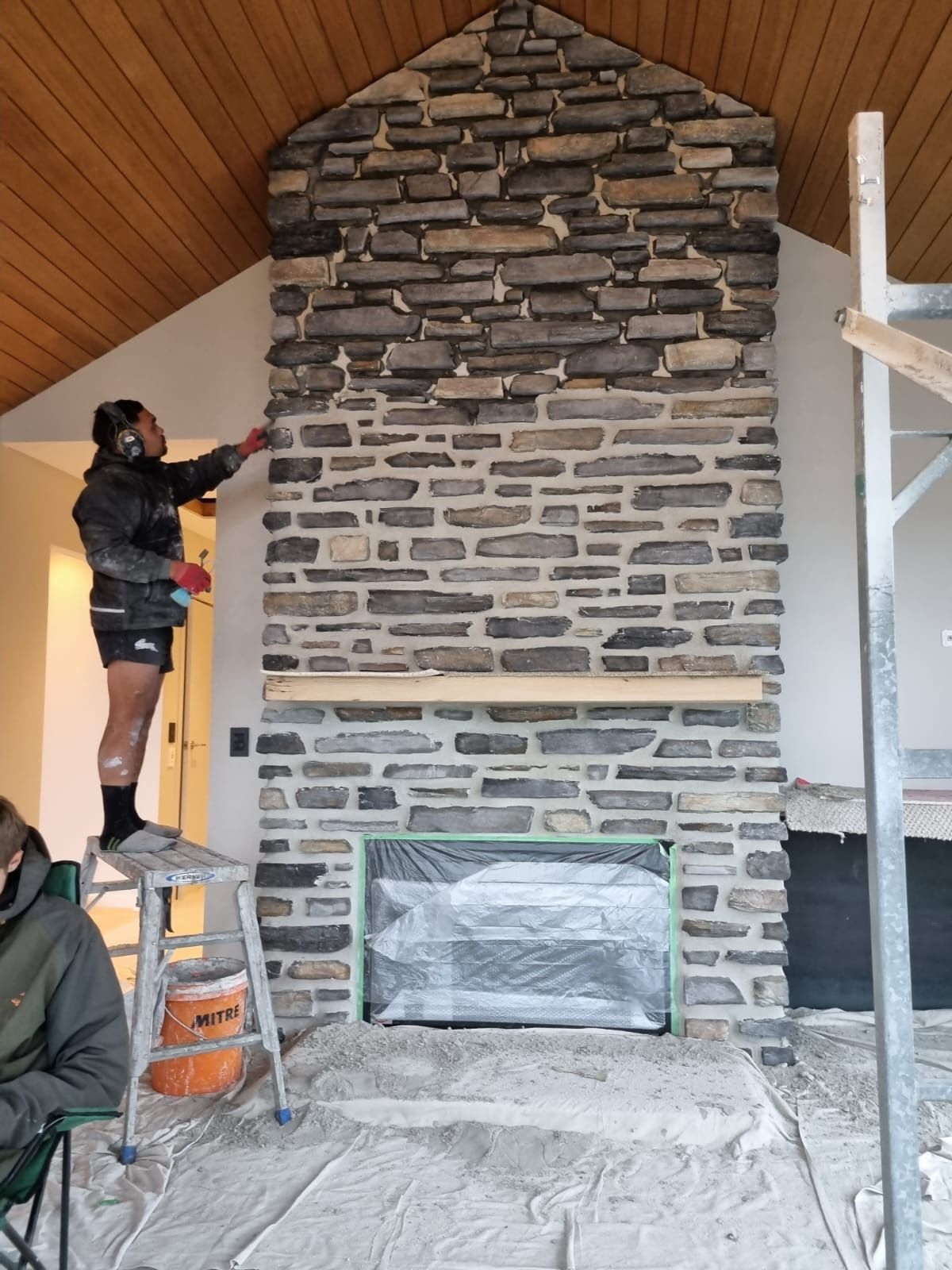 A man is standing on a ladder painting a stone fireplace.