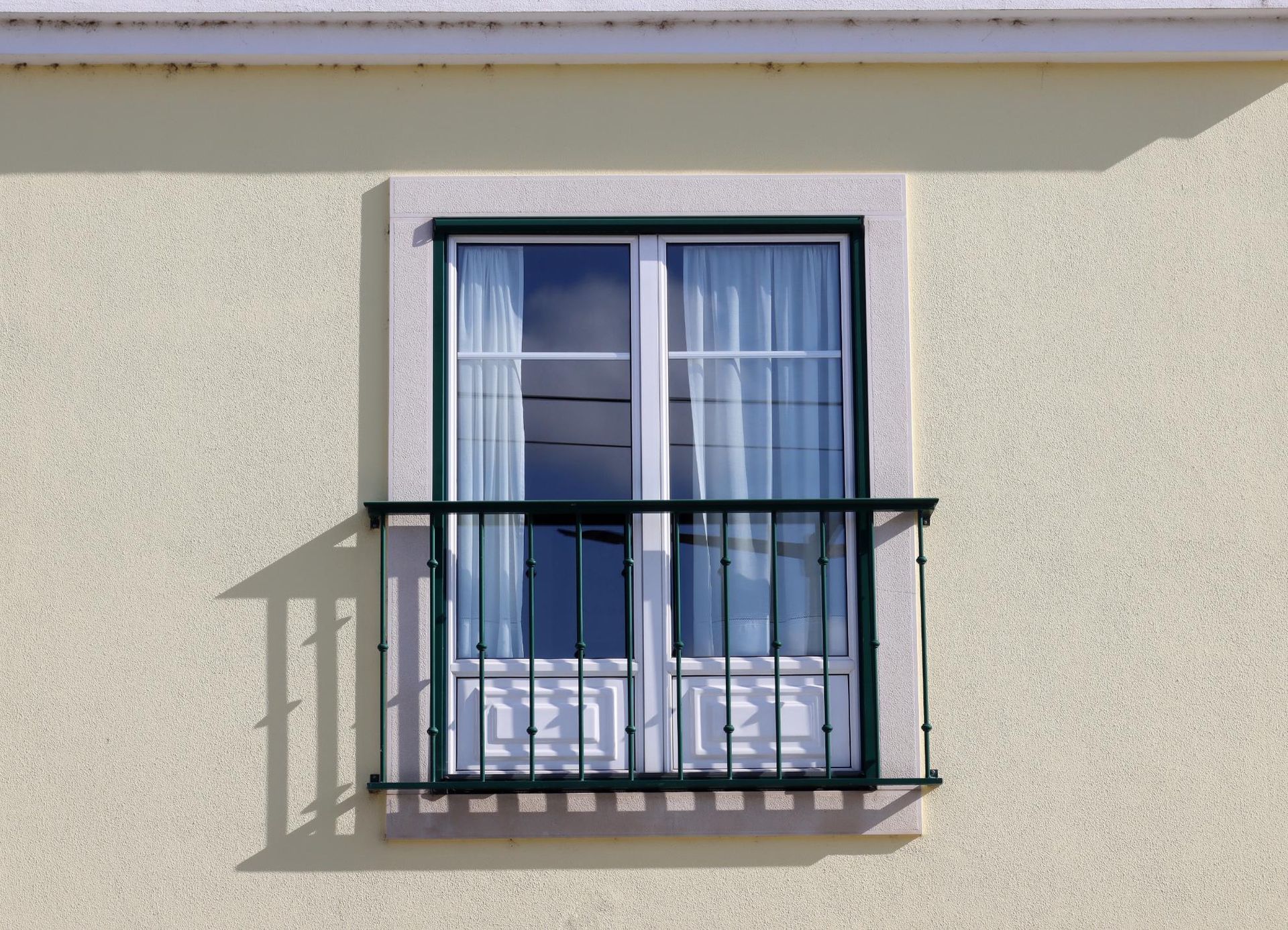Window with white trim, green railing, and sheer curtains on a stucco building, casting a shadow.