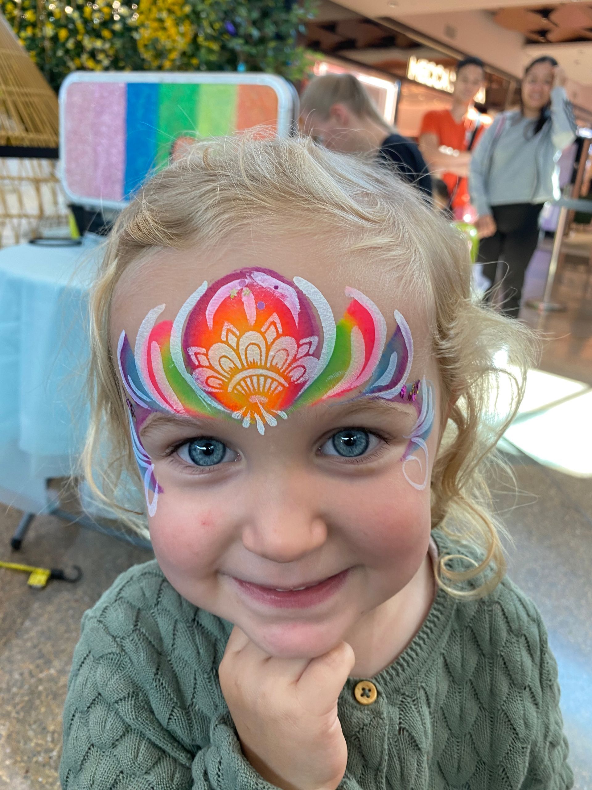 Young child with blue eyes and face painted with rainbow flower design, smiling.