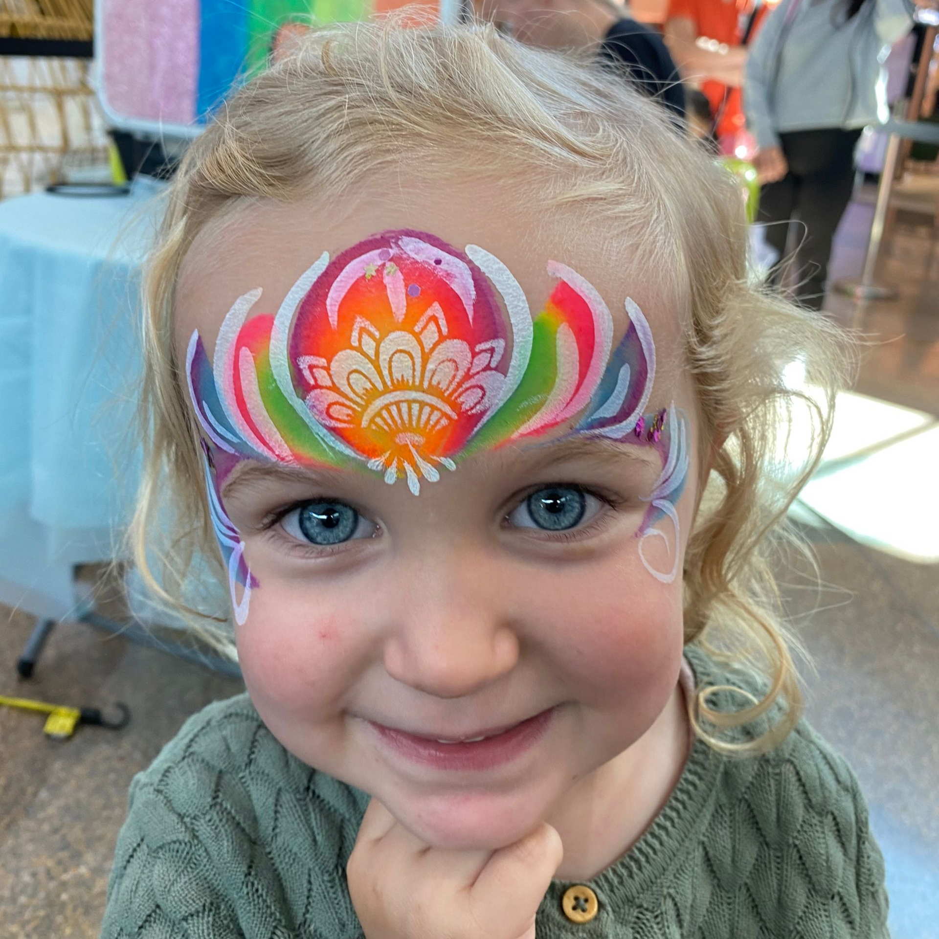 Girl with blue eyes smiles, face painted with a rainbow flower design at an event.
