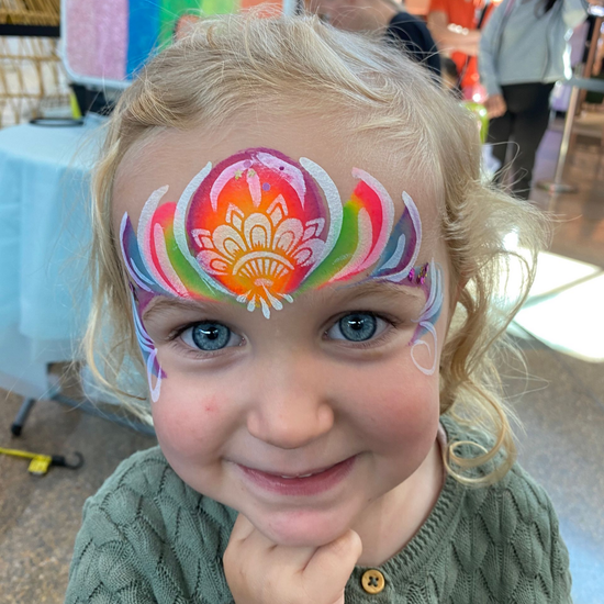 Girl with blue eyes smiles, face painted with a rainbow flower design at an event.