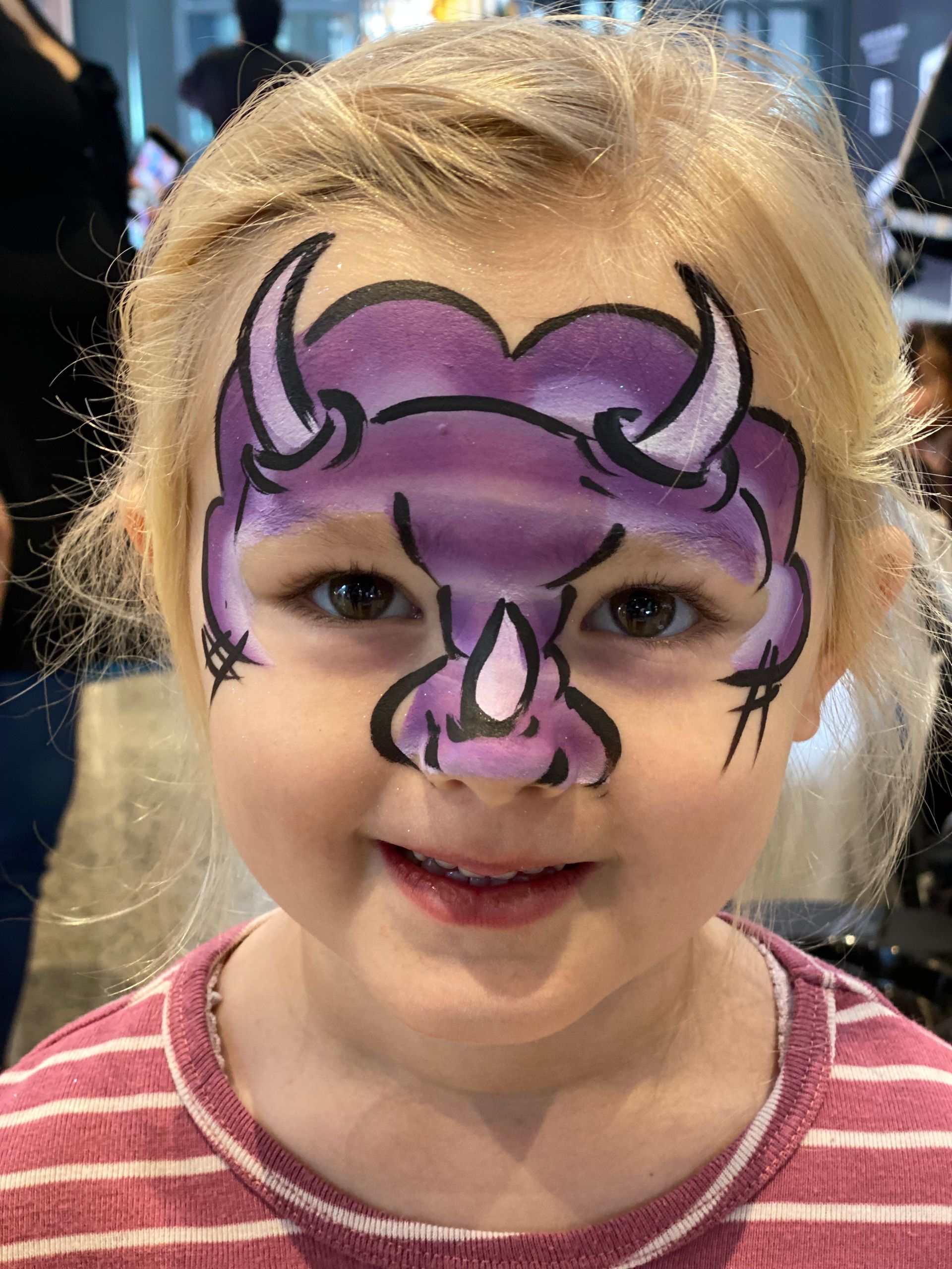 Young girl with purple dinosaur face paint, smiling.