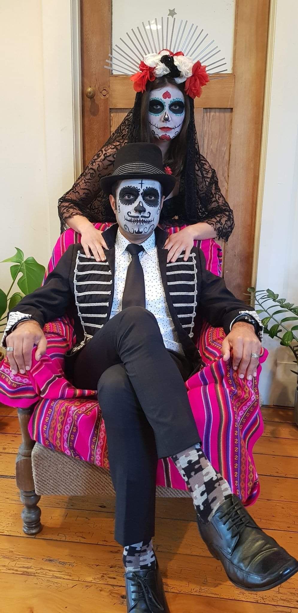 Couple in Día de Muertos makeup and costumes, posing. Man in a chair, woman behind.