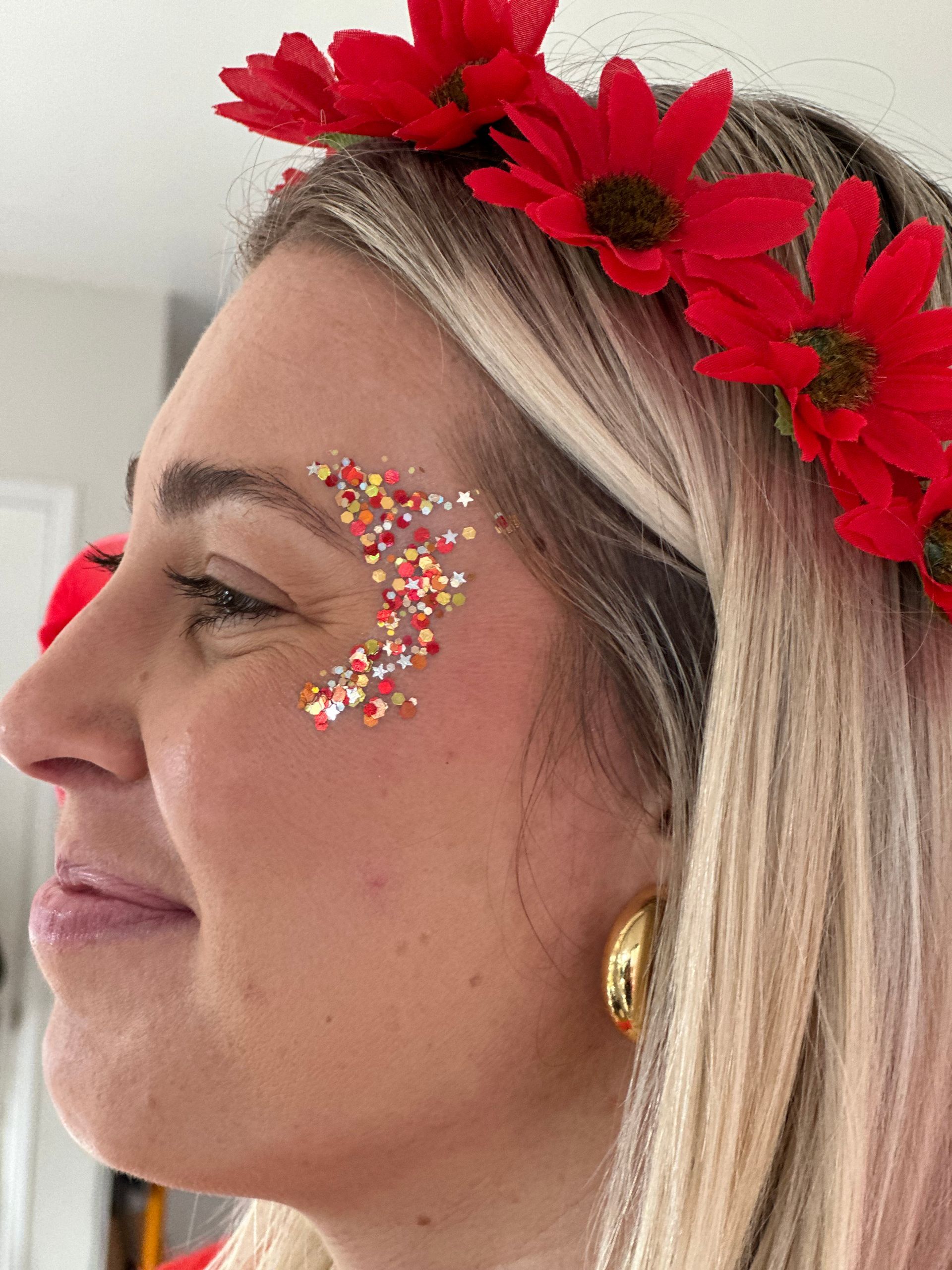 Woman with red flower crown, glitter on cheek, and gold earrings.