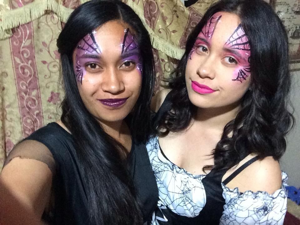 Two women in Halloween costumes with spiderweb face paint, smiling.