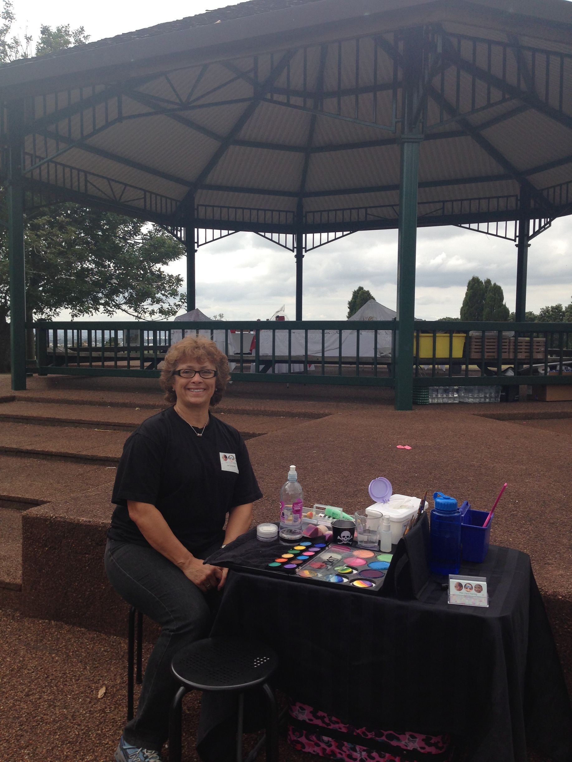 Woman at a table with face paint supplies, in front of a gazebo.