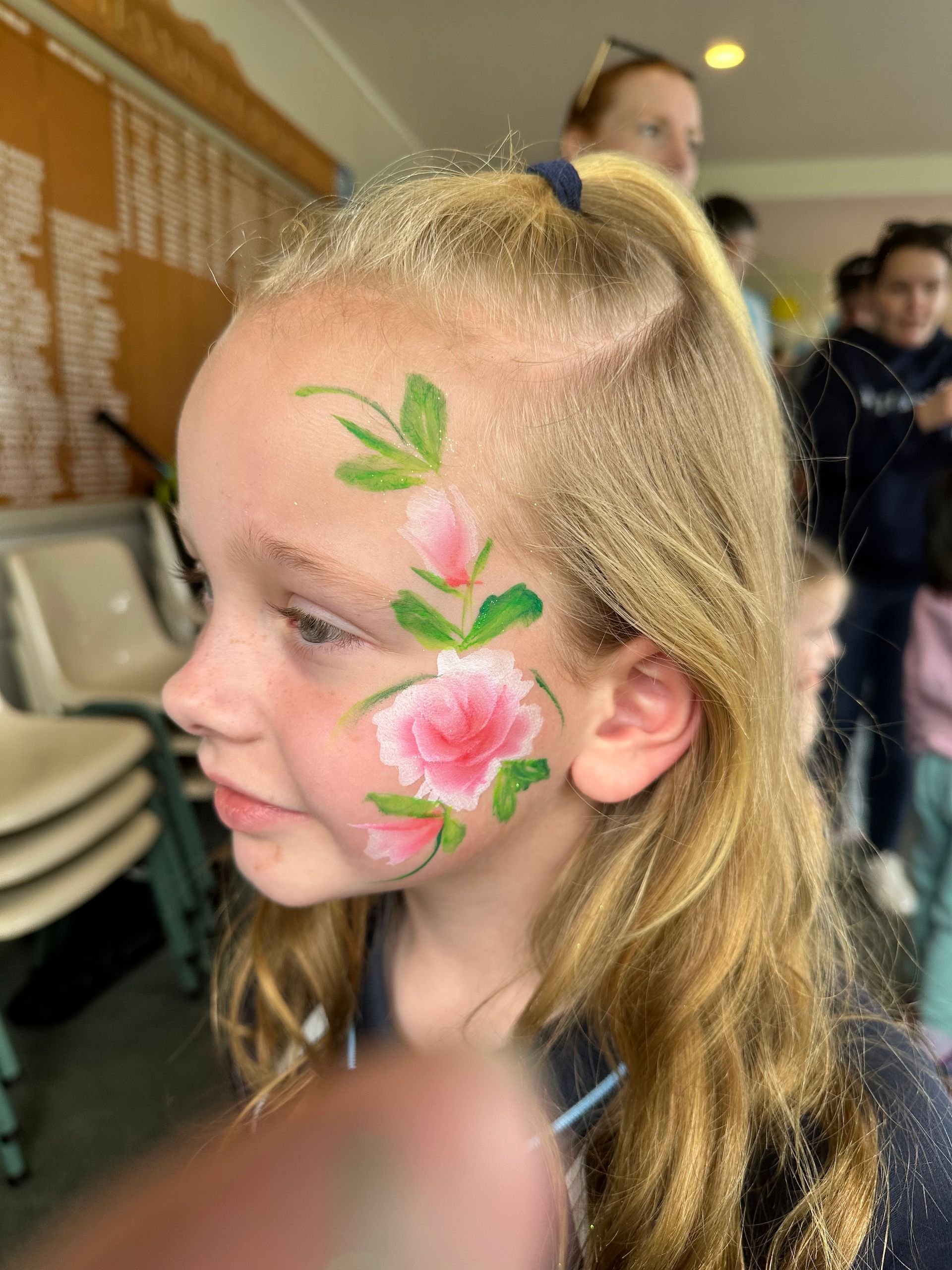 Girl with flower face paint; pink blossoms, green leaves on her cheek, blonde hair, smiling slightly.