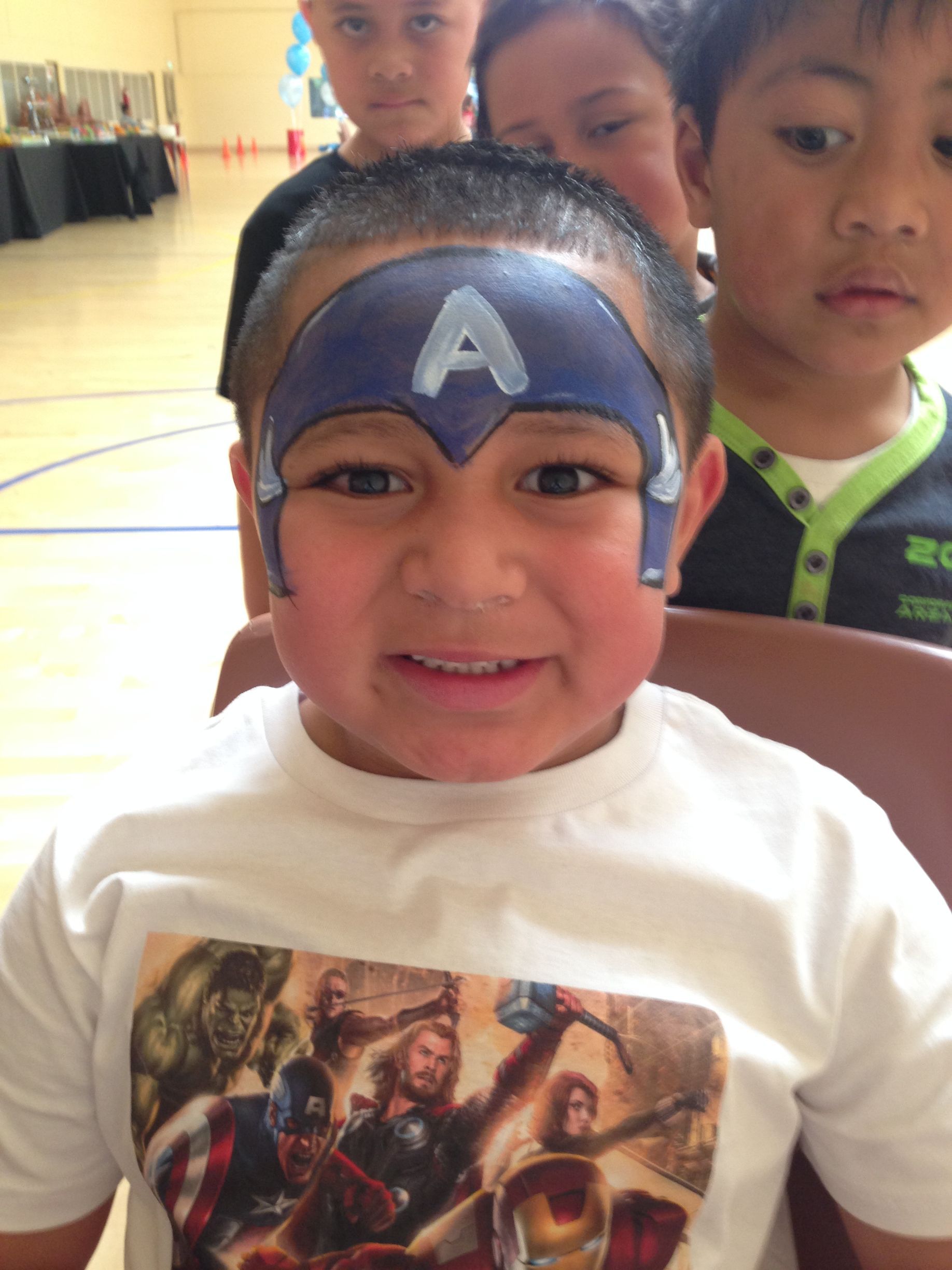 Boy with Captain America face paint, wearing a shirt with Avengers characters, smiling.