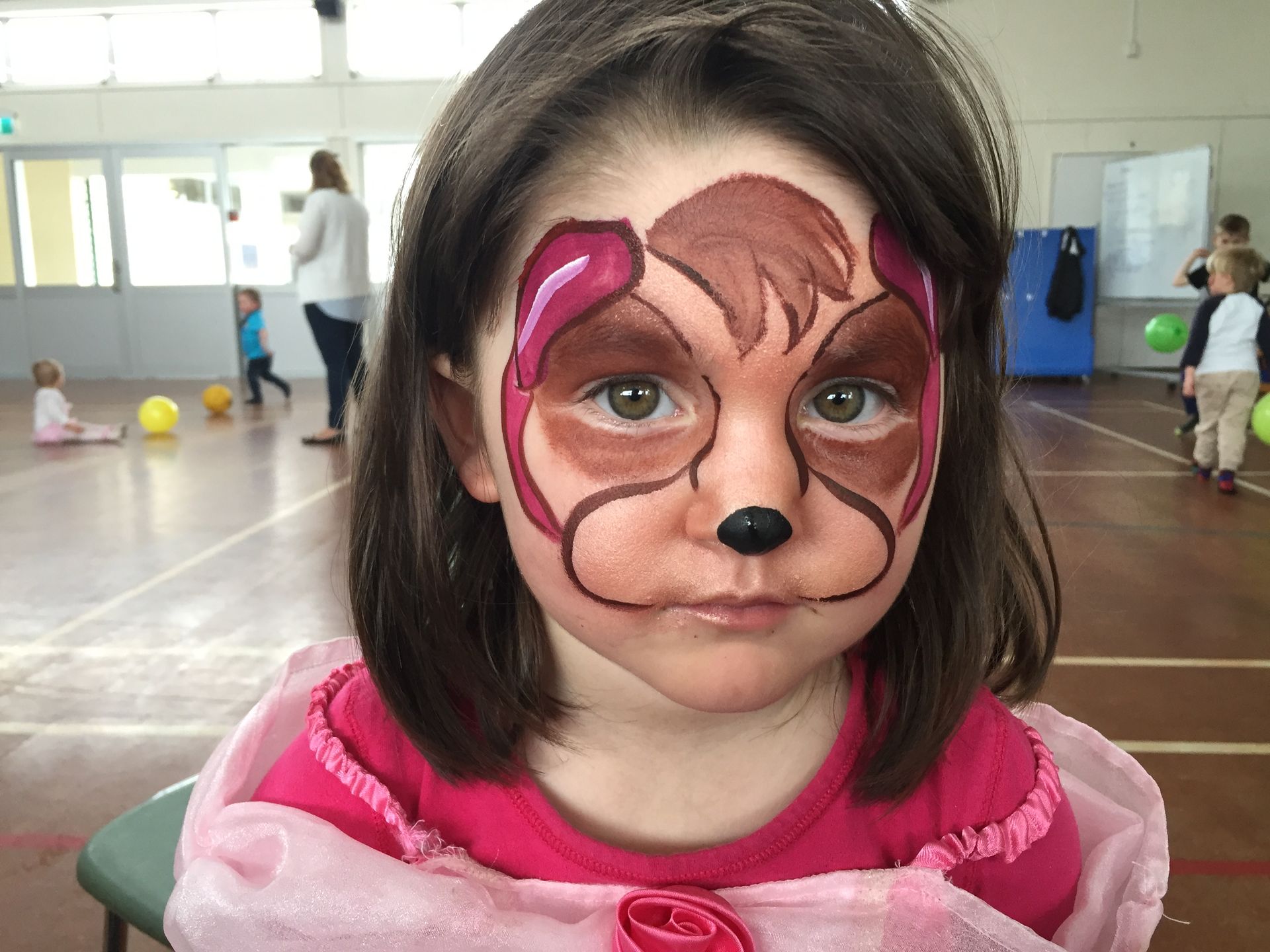 Girl with dog face paint; brown, pink colors. Indoor gym setting.