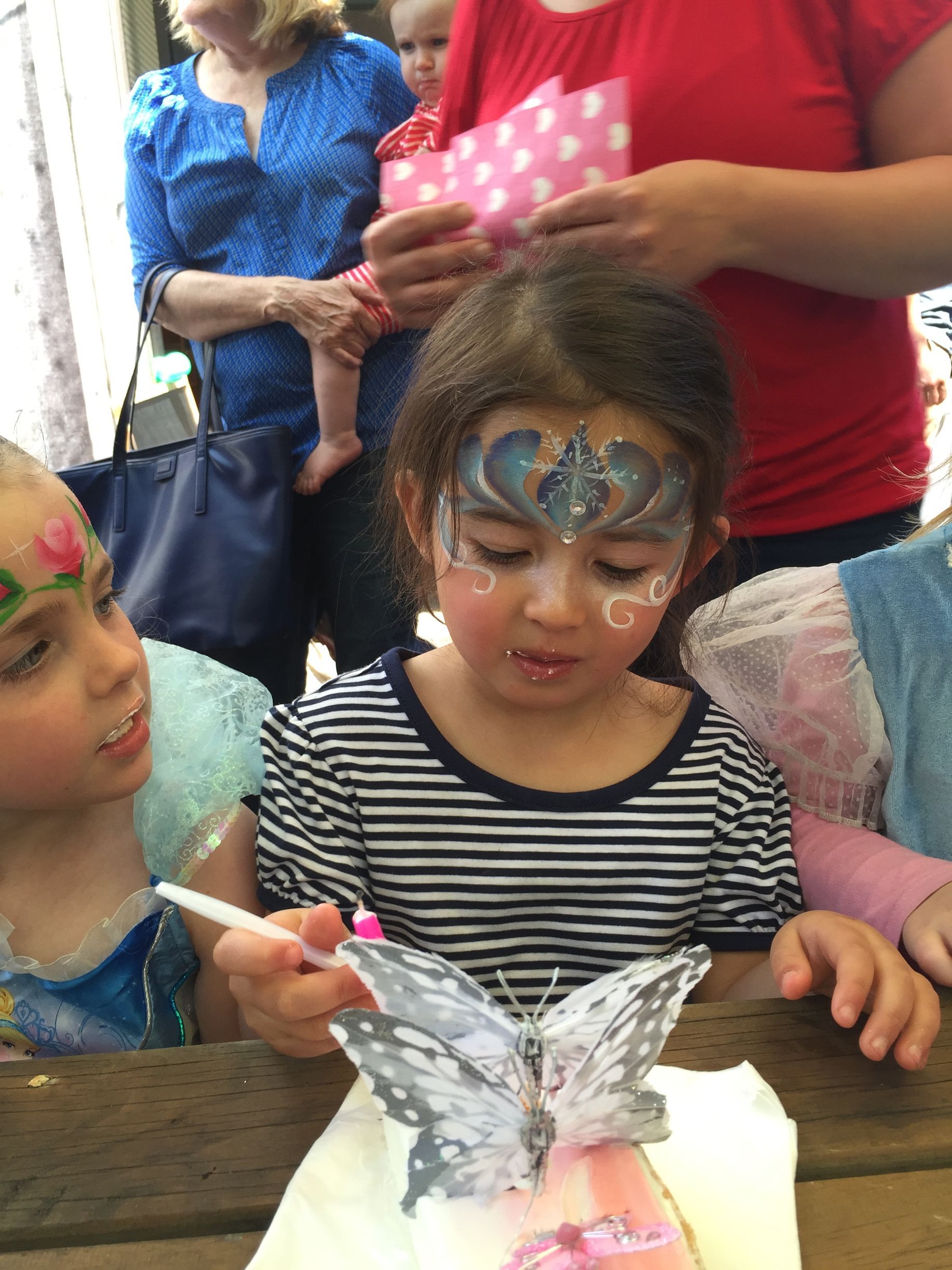 Girl with face paint holding a butterfly, other children and adults in the background.