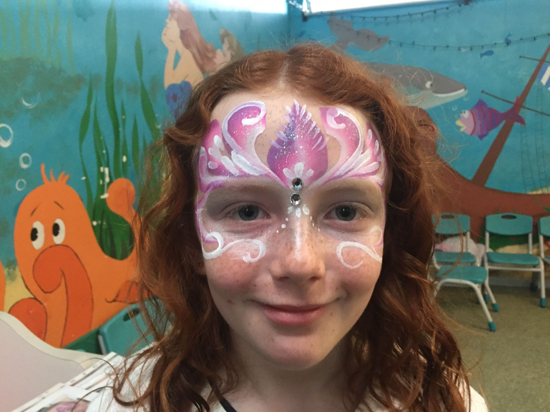 Girl with red hair, face painted with a pink and white floral design, in front of a mural.