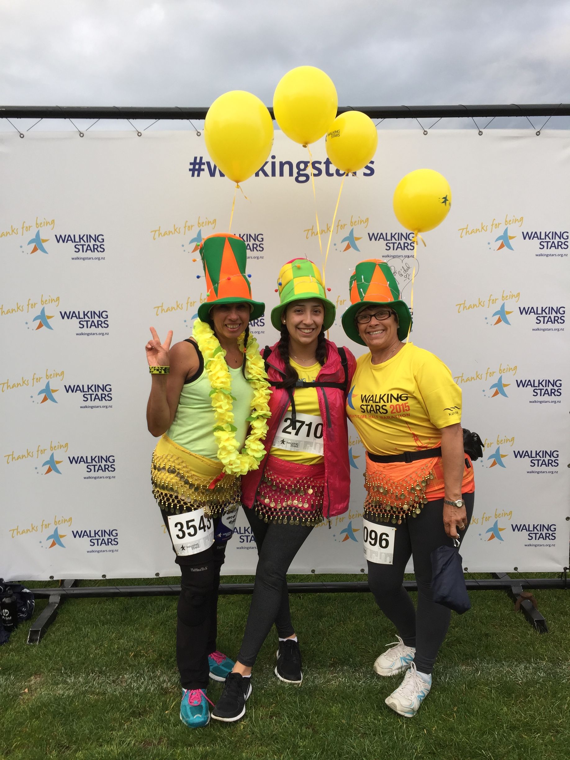 Three women in colorful costumes pose with yellow balloons at an outdoor event, smiling and giving peace signs.