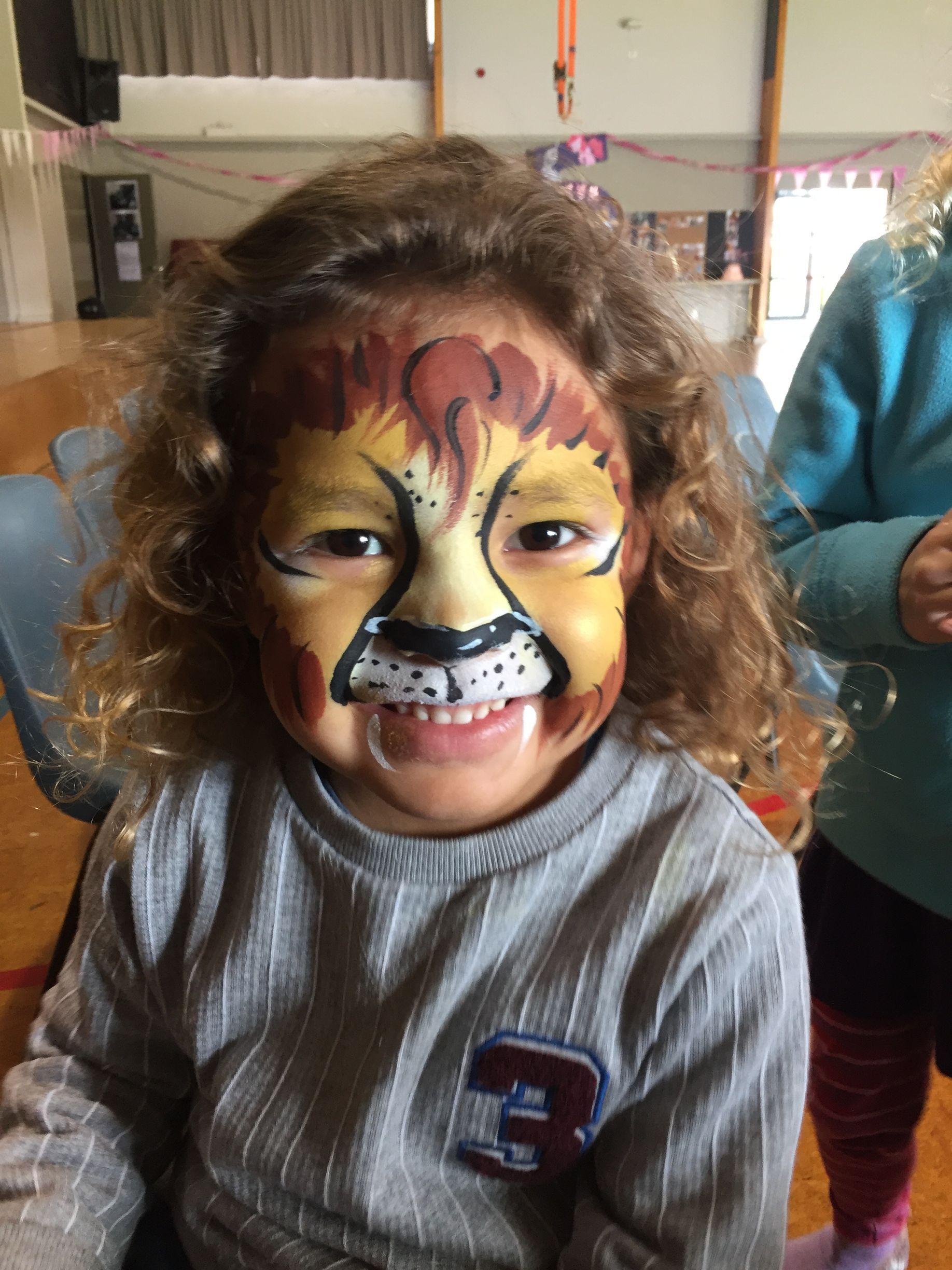 Child with lion face paint smiling. Indoors, wearing a gray shirt.